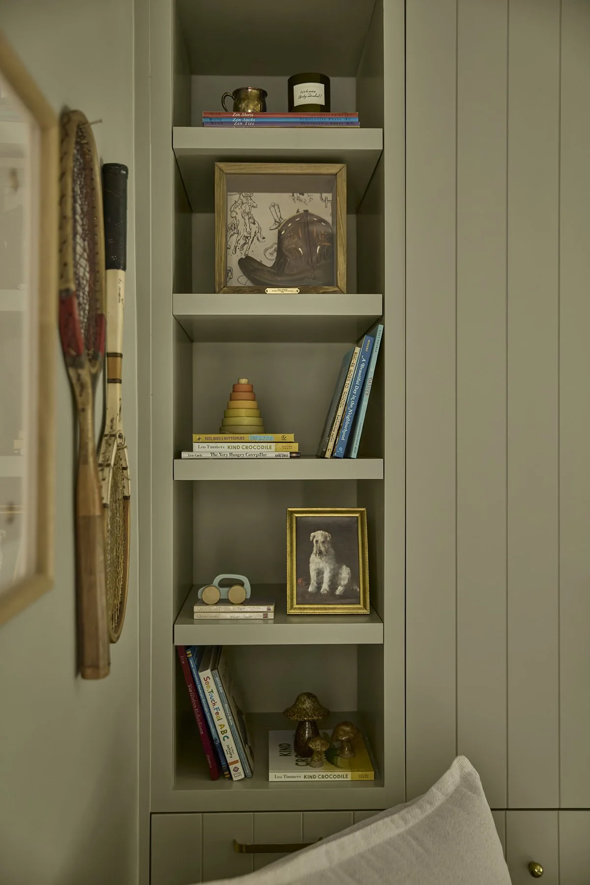 A built-in bookshelf with four shelves inside a room. The top shelf holds a small metal pitcher, a black and white candle, and a row of books. The second shelf contains a framed picture of a baseball glove and ball. The third shelf has a stack of col