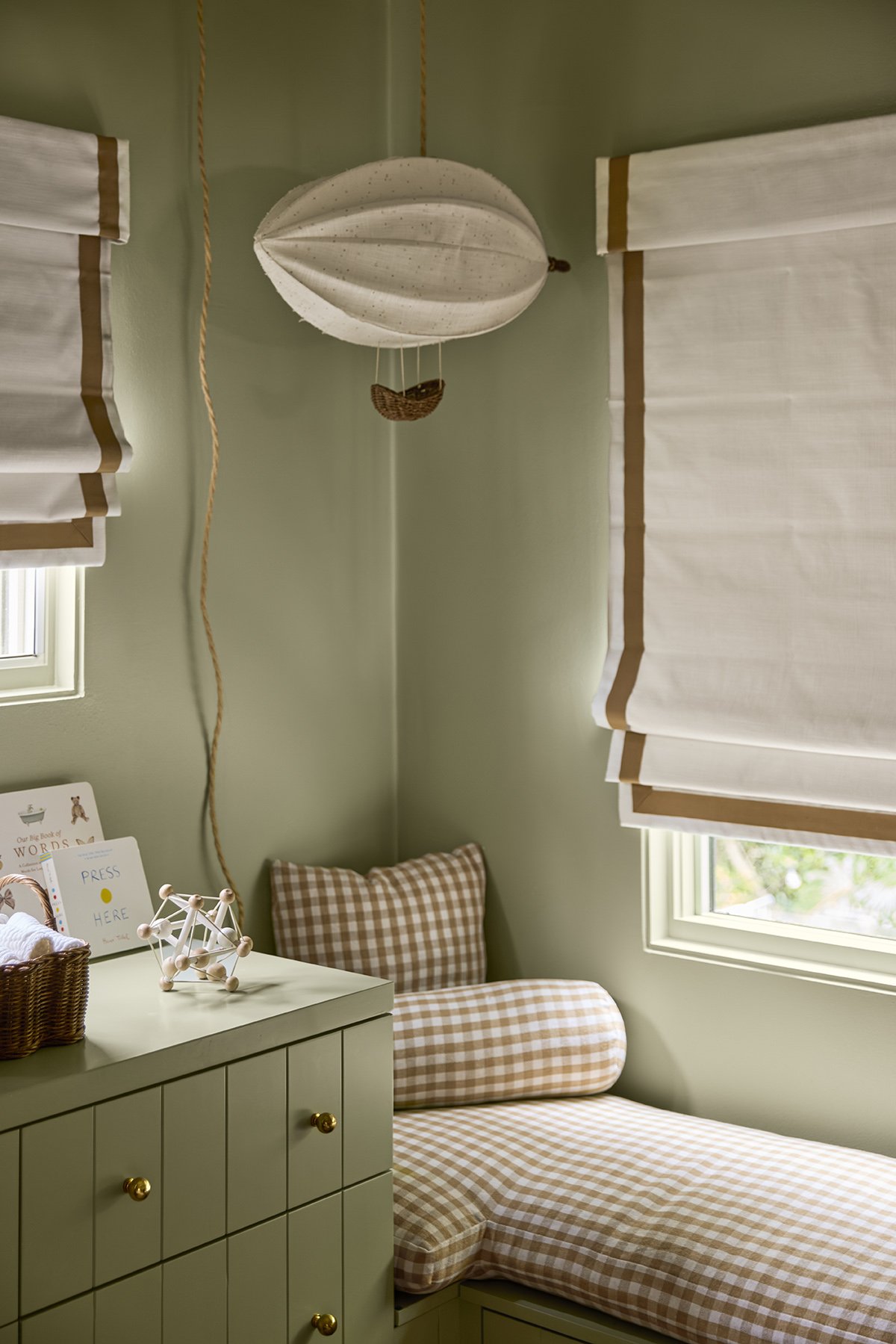 A cozy bedroom corner with a beige checkered bed and matching headrest, a light green dresser with a small sign, woven basket, and decorative molecule sculpture, green walls, and windows with white curtains.