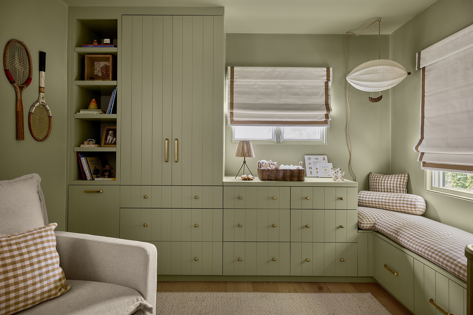 nursery room with mint green painted cupboards and drawers. Decorative tennis wrackets are hanging on the wall above a beige armchair with a light brown gingham patterned pillow. Windows are decorated with warm white shoji blinds. Under one window is
