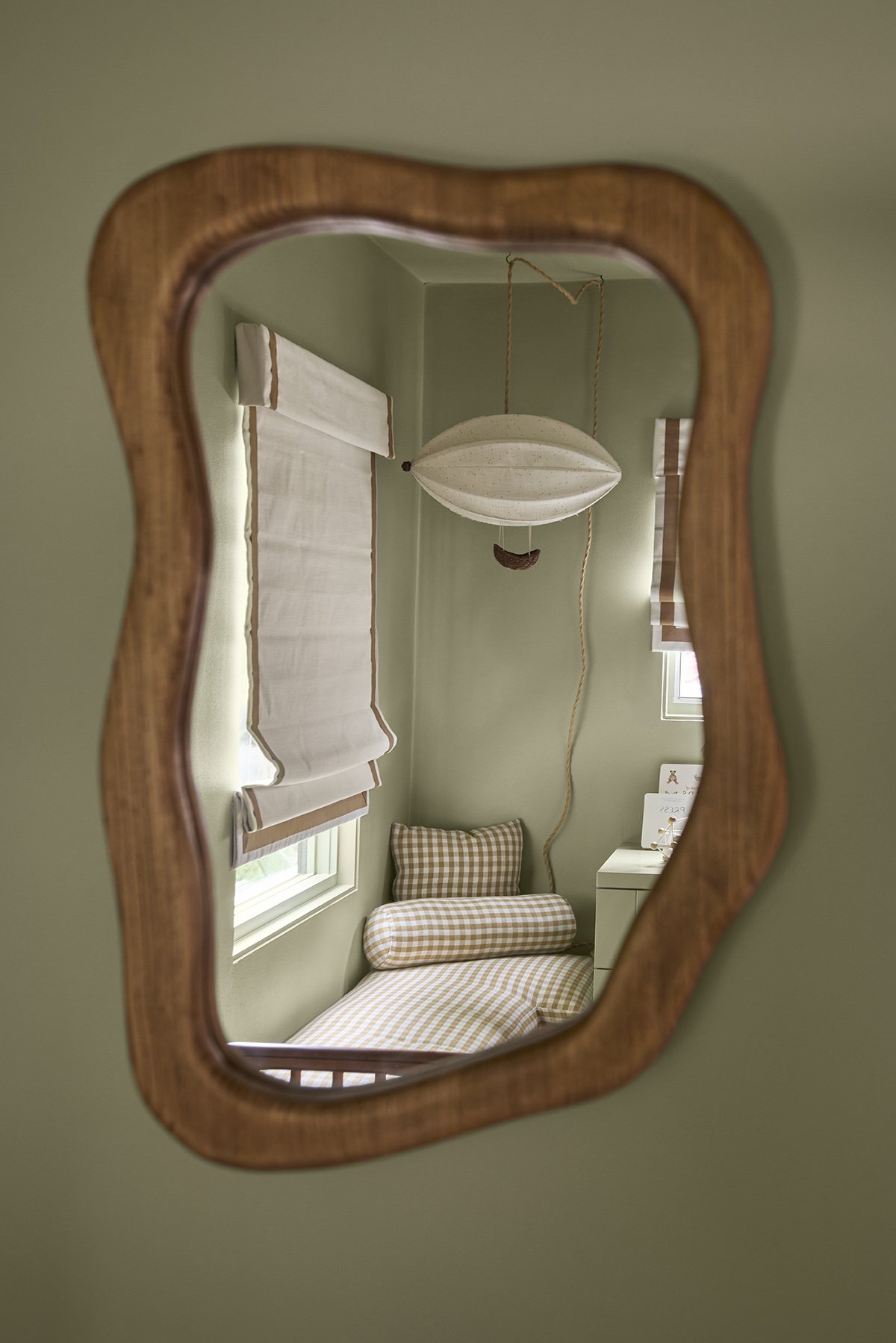 A room with green walls, a window with white curtains, a checkered bed with a matching pillow and bolster, a small white dresser, and a decorative hanging lamp; viewed through a wooden-framed mirror with a wavy edge.