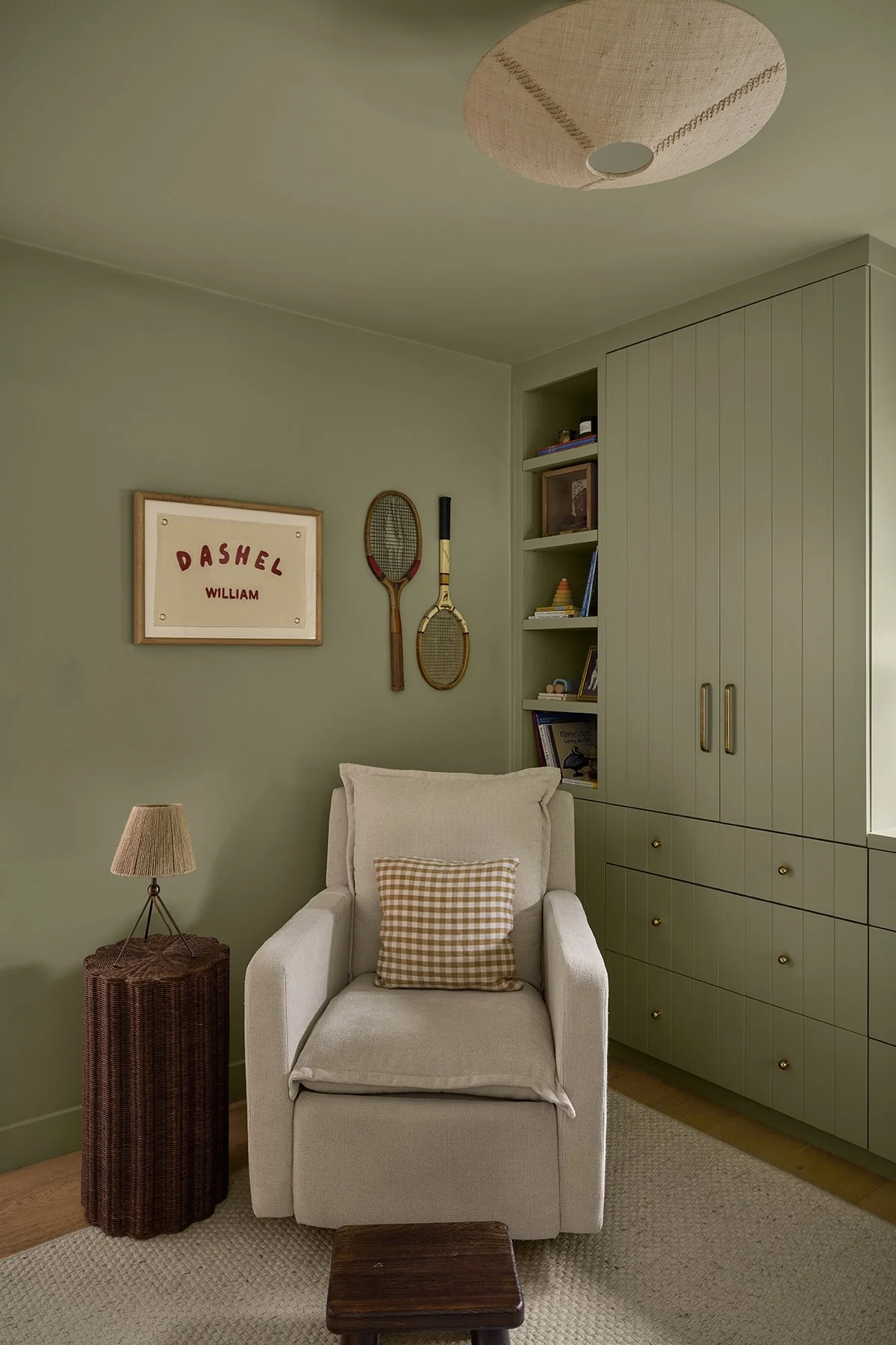 Cozy living room corner with a beige armchair, a checkered pillow, a wicker side table, a floor lamp, wall-mounted tennis rackets, a framed sign reading "Dashel William," a bookshelf with books and decorative items, and a large light fixture on the c