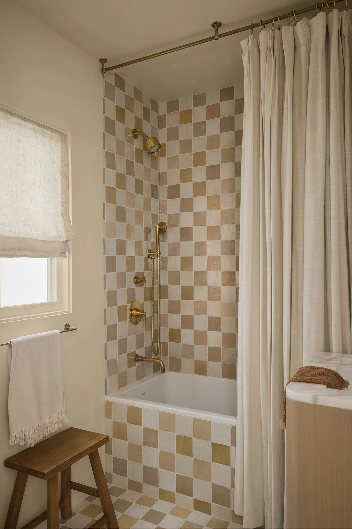 Bathroom shower with a checkered tile wall in beige, tan, and cream, gold fixtures, a small window with a white curtain, a towel bar with a white towel, a wooden stool, and beige curtains dividing the shower area from the rest of the bathroom.