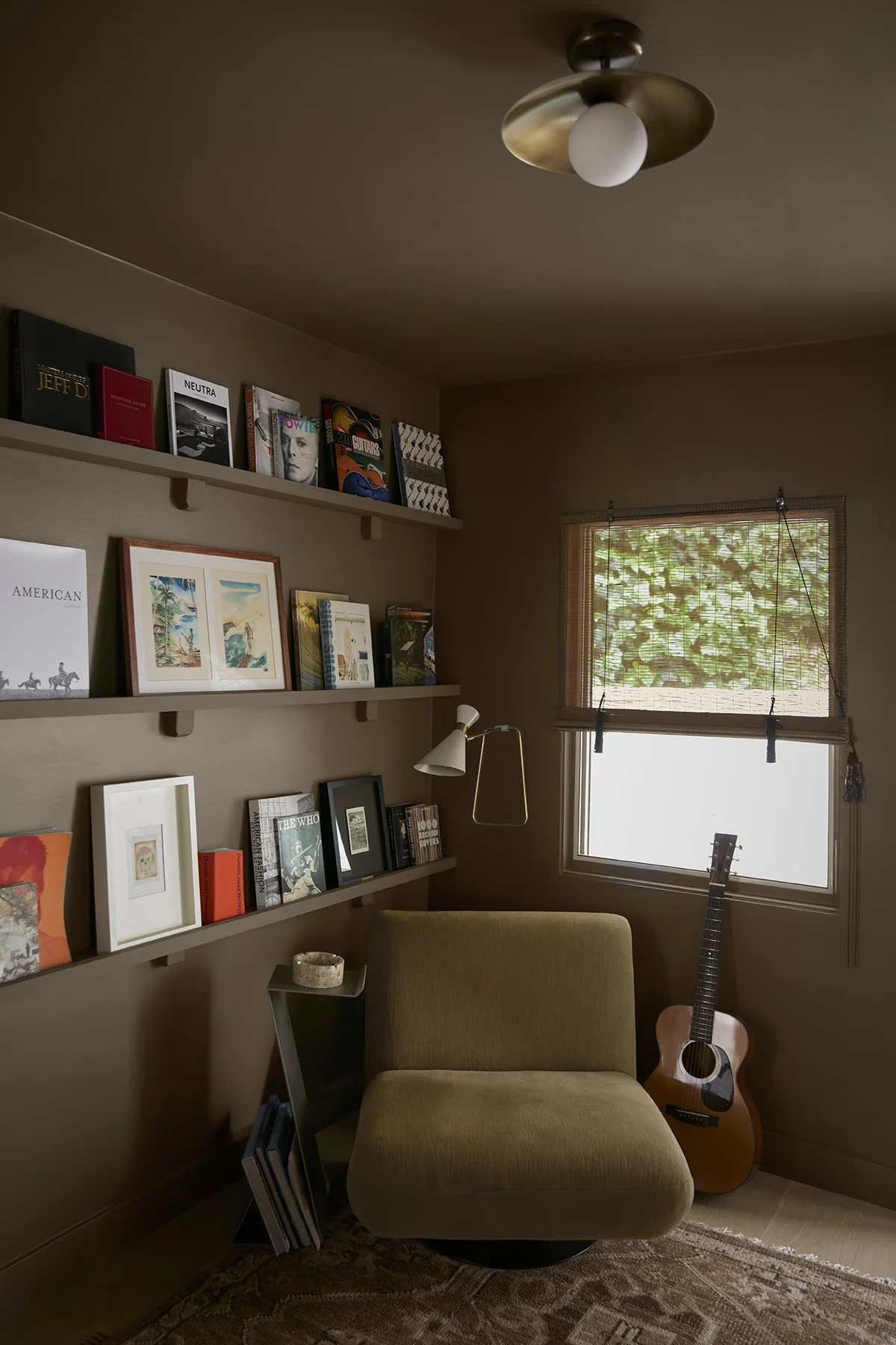 A cozy room with beige walls and a window covered by a woven shade. There are three floating shelves filled with books and framed pictures. A small side table with a decorative bowl stands next to a green upholstered armchair. A guitar leans against 
