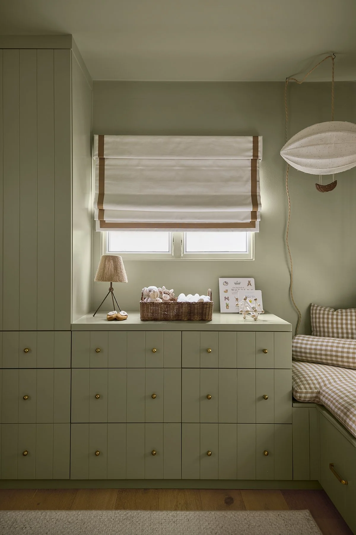 A cozy nursery with green built-in cabinets featuring gold knobs, a window with white shades, a wicker basket with plush toys, a small table lamp, and display boards with art and decorations. There's a checkered armchair on the right side.
