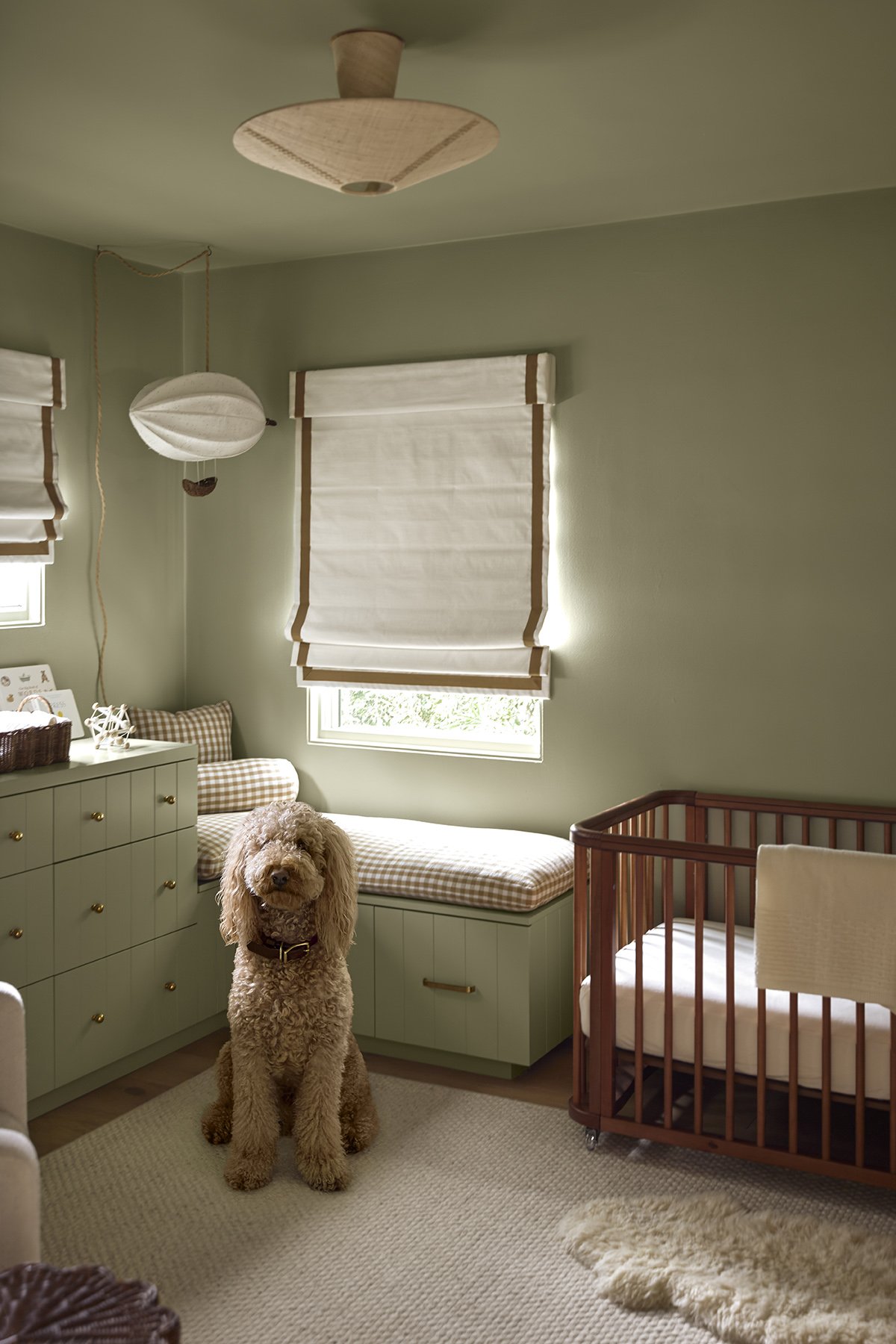 A cozy nursery with light green walls, white window shades, a cream carpet, and a green dresser with gold knobs. There is a plaid cushioned window seat, a brown wooden crib, and a fluffy beige rug. A large, curly-haired dog sits in the middle of the 