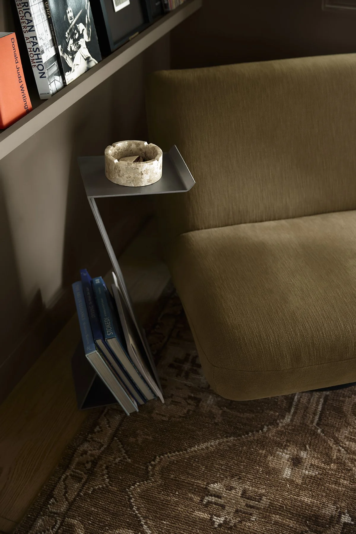 A beige upholstered armchair next to a small black bookshelf containing several books, with a small gray side table beside it. On the side table, there is a round ashtray, and above the armchair, a shelf with more books is visible.