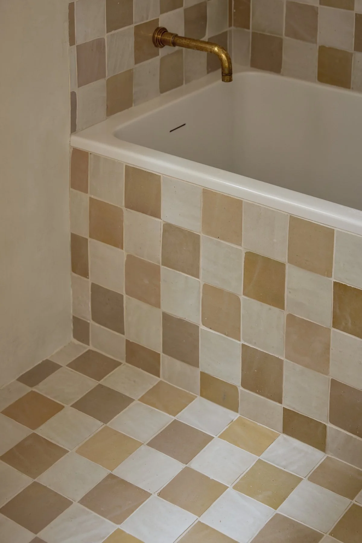 Close-up of a bathroom corner with beige and brown checkered tiled walls and floor, featuring a white bathtub with a brass faucet.