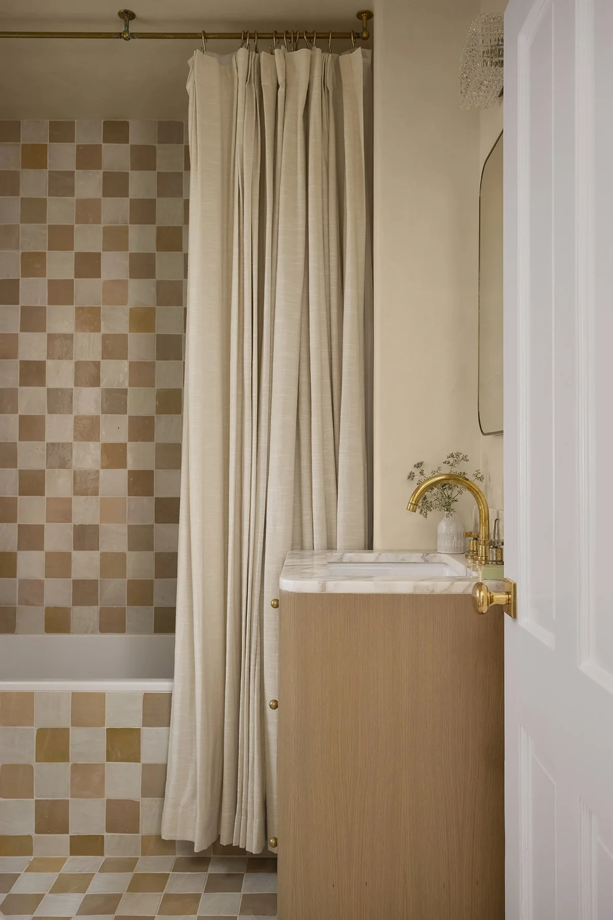 A bathroom with a shower area that has a curtain and a wall with beige, pink, and white checkered tiles, a small vanity with a marble top, a gold faucet, and a mirror on the wall.