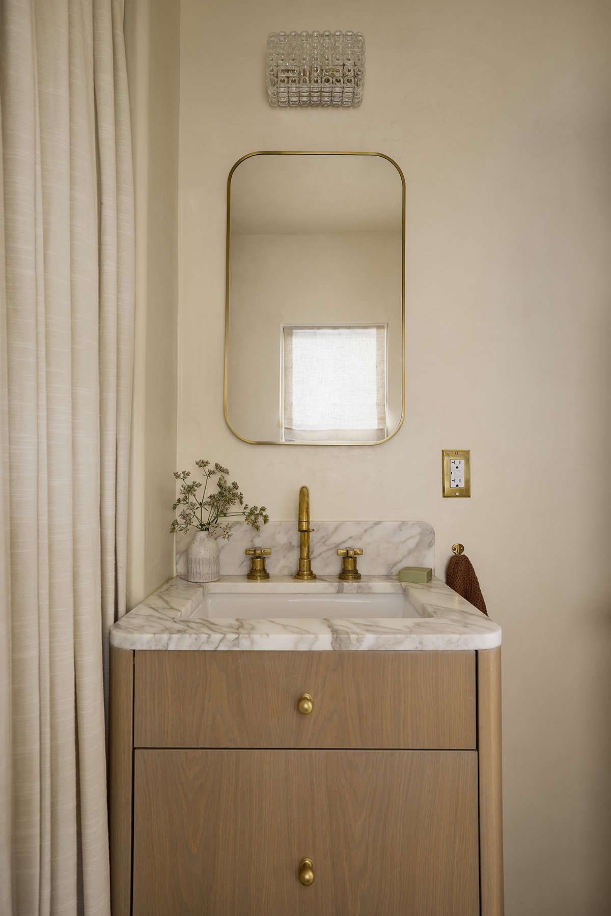 A bathroom vanity with a wooden cabinet, white marble countertop, and gold fixtures. A mirror with a gold frame hangs above, and a small vase with dried flowers is on the left. To the right, there is a bar of soap, a brown towel, and a beige wall wit