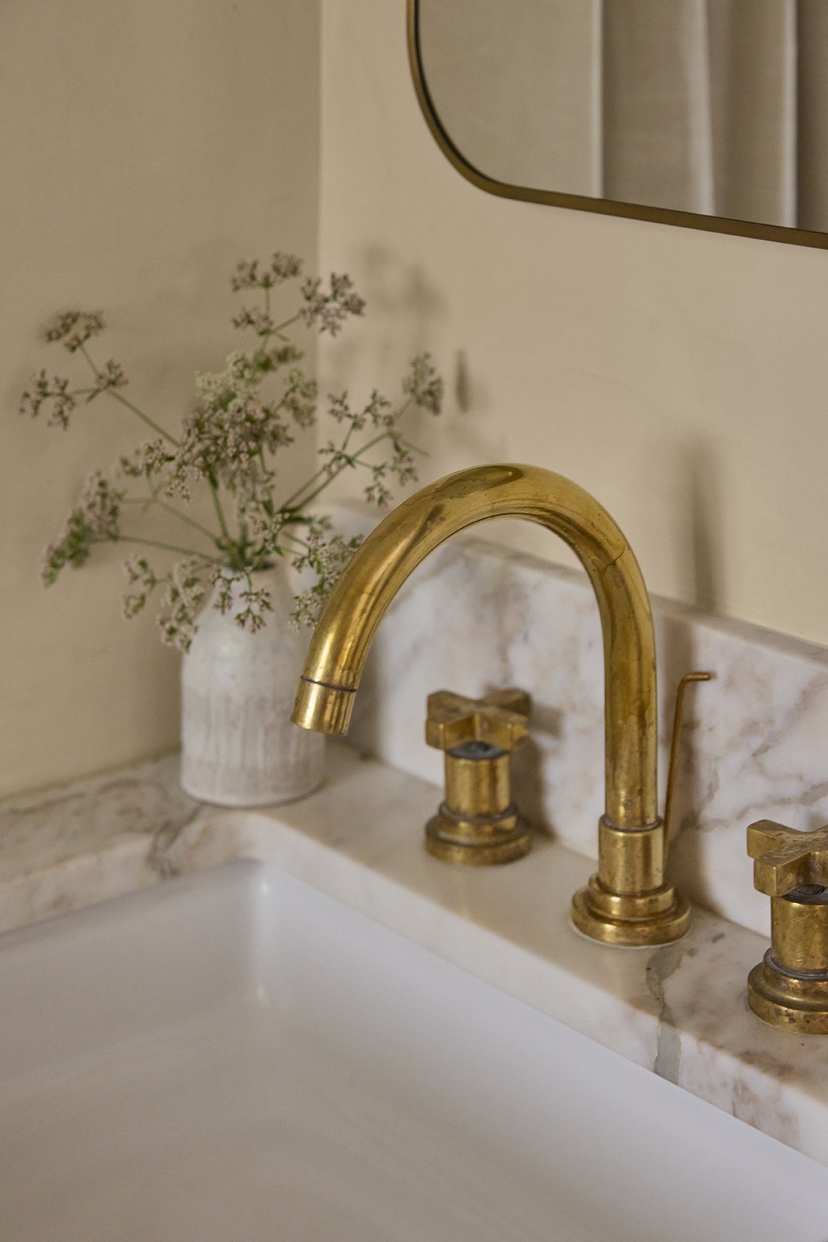 A bathroom sink with a gold-colored faucet, a white marble countertop, a mirror, and a white vase with flowers.