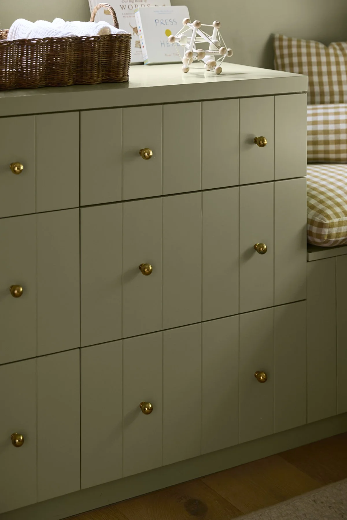 A light green cabinet with gold knobs in a room corner, with a basket and books on top and a checkered cushion on the side.