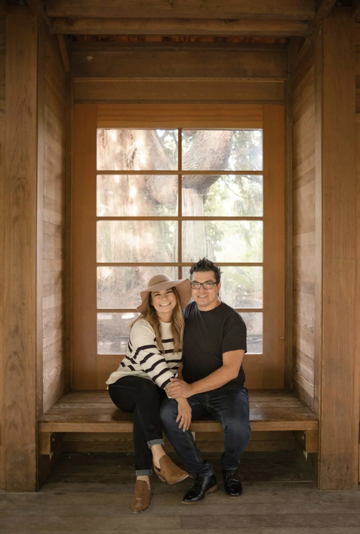 A smiling couple sitting on a wooden bench in front of a large window with a view of trees outside. The woman is wearing a tan hat, a white and black striped sweater, and tan shoes. The man is wearing glasses, a black t-shirt, and black shoes.