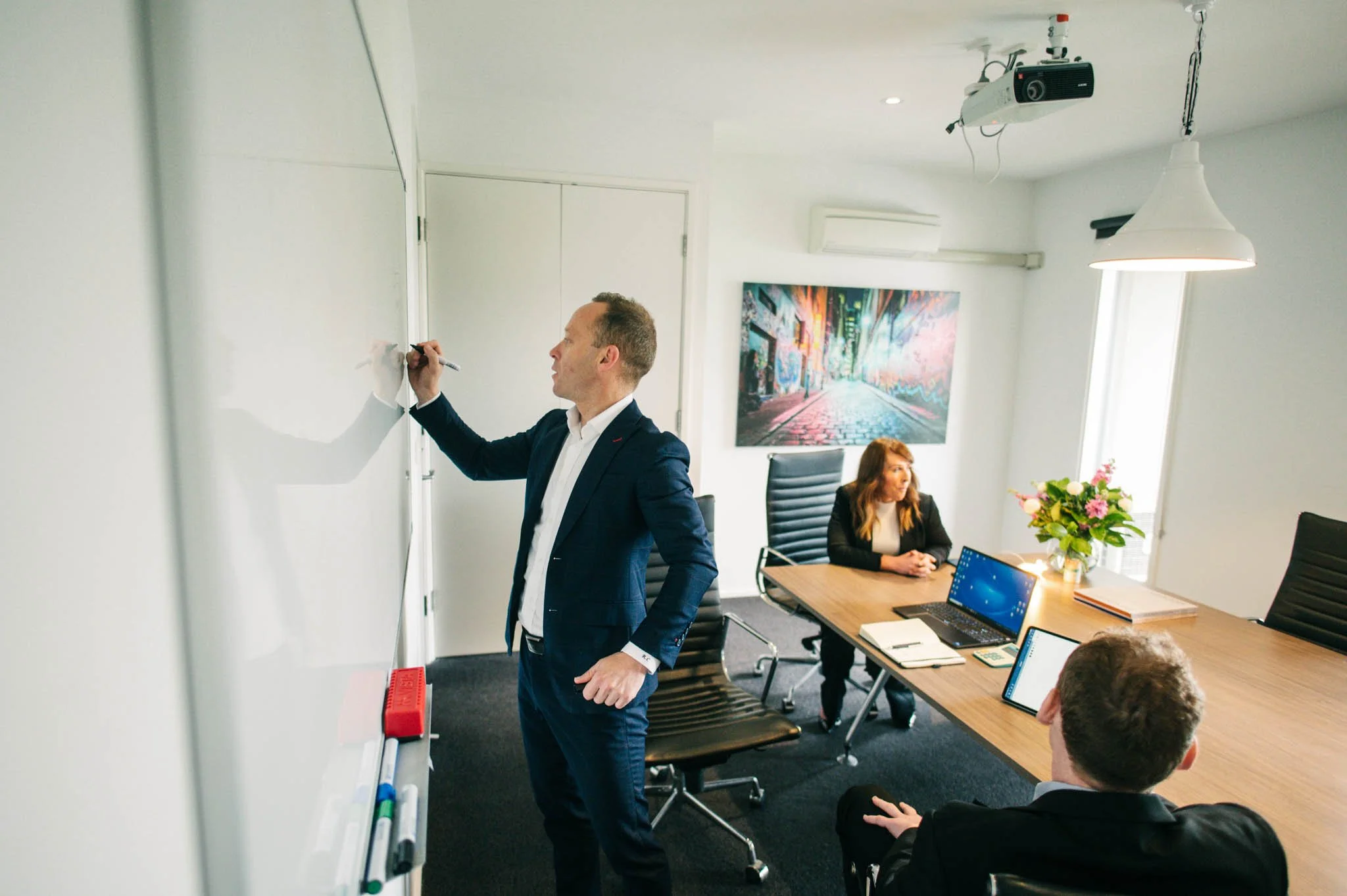 Man in a suit writing on a whiteboard in a conference room with two women seated at a table, one of whom is using a laptop, and a large colorful cityscape artwork on the wall.