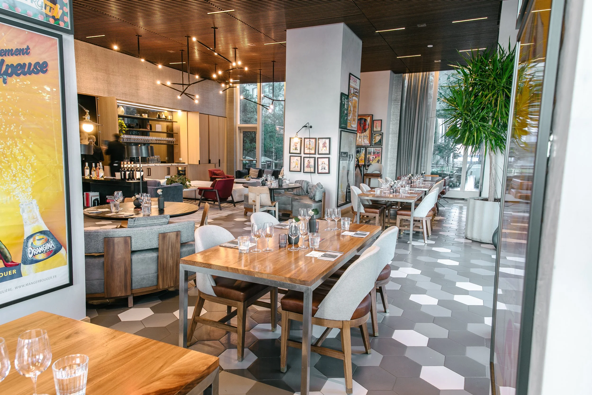 Interior shot of One Manhattan Square dining room showing a bar, lounge area, and wooden tables set with glasses, surrounded by floor-to-ceiling windows.