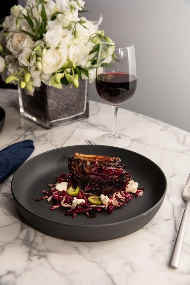 A plated dish featuring roasted radicchio, crumbled cheese, and green garnish on a black plate, with a glass of red wine.