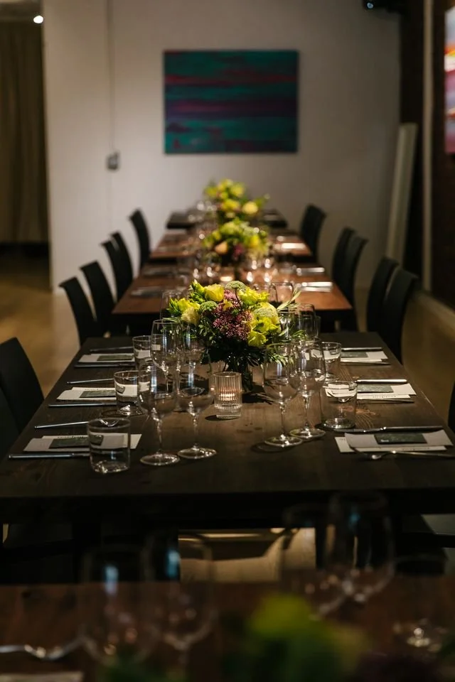 A long dinner table dressed for a special event with polished glassware, white plates, folded napkins, and a lush floral arrangement.