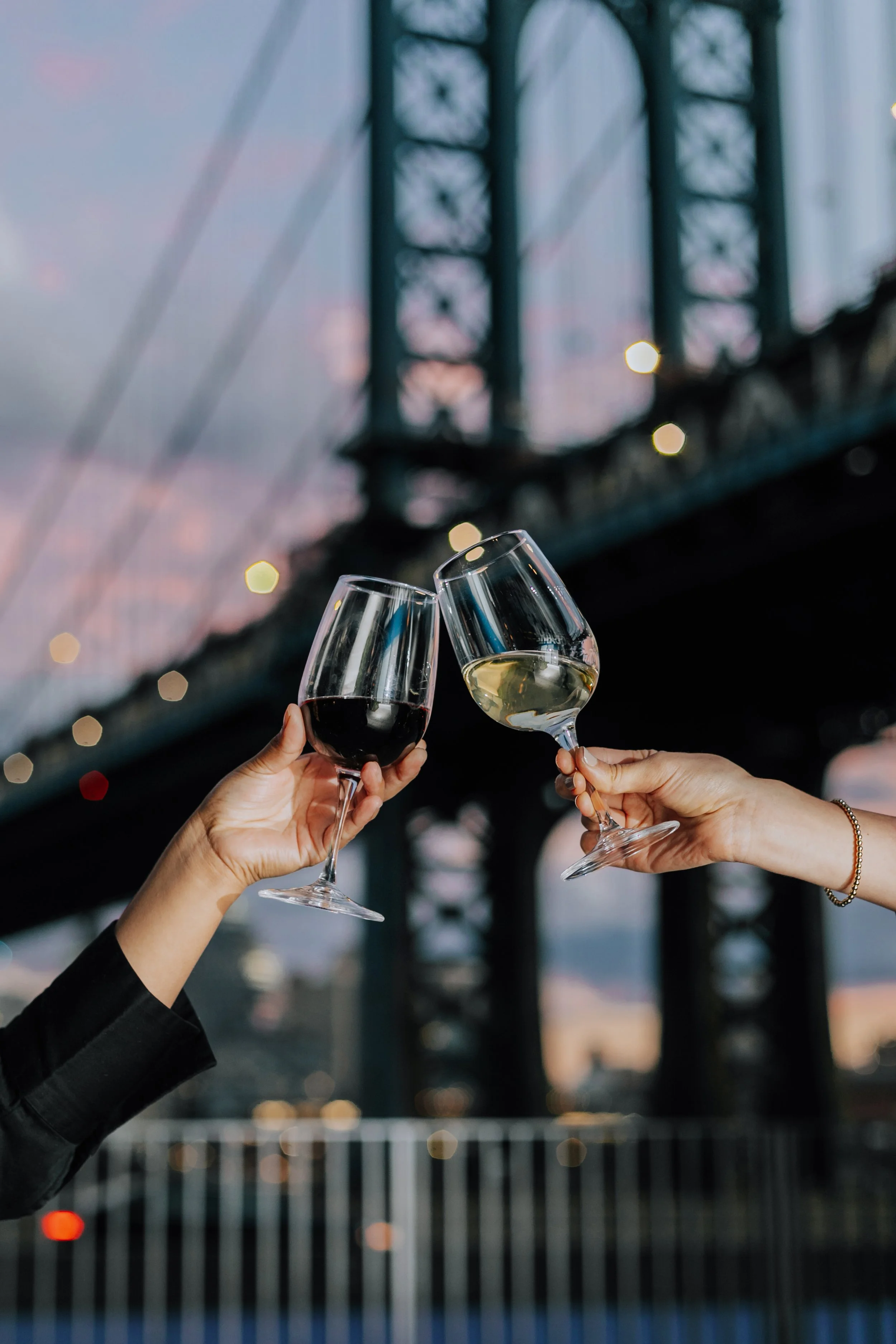Two cropped arms holding wine glasses in a toast in front of a blurry backdrop of the Brooklyn Bridge at dusk.