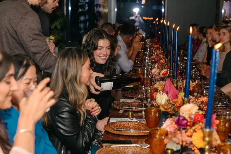 Guests seated at a long dinner table as wine is poured, with blue taper candles and floral centerpieces.
