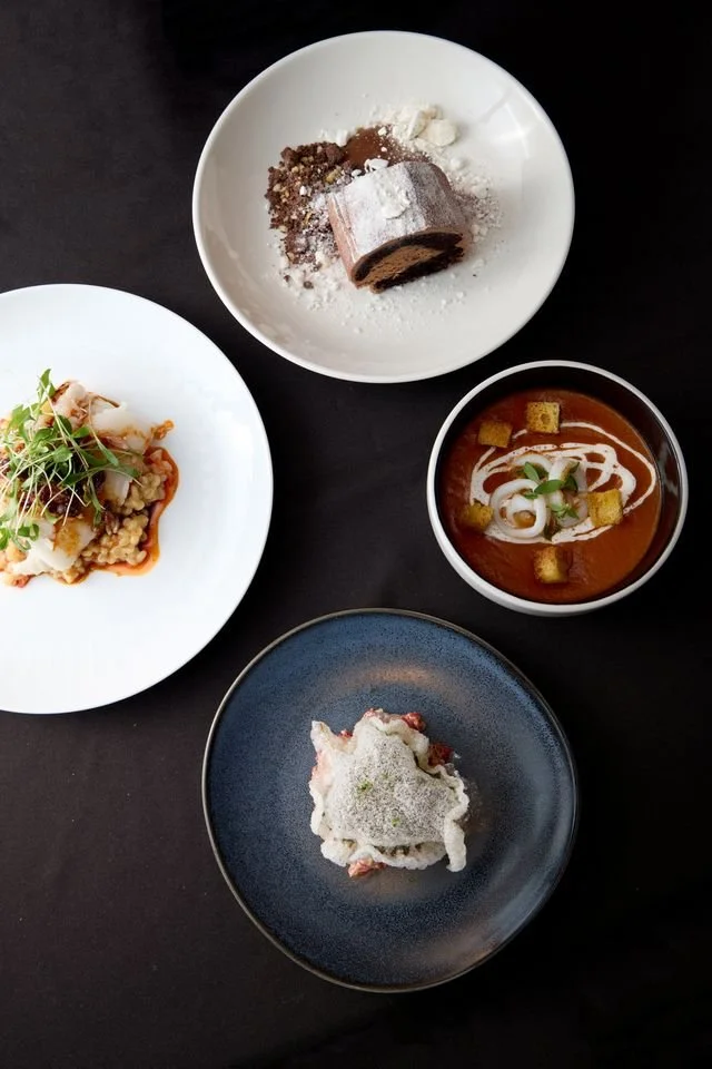 Aerial view of four elegant dishes on a dark tablecloth: a chocolate dessert with powdered sugar, tomato soup with croutons, a savory entree with greens, and a delicate appetizer.