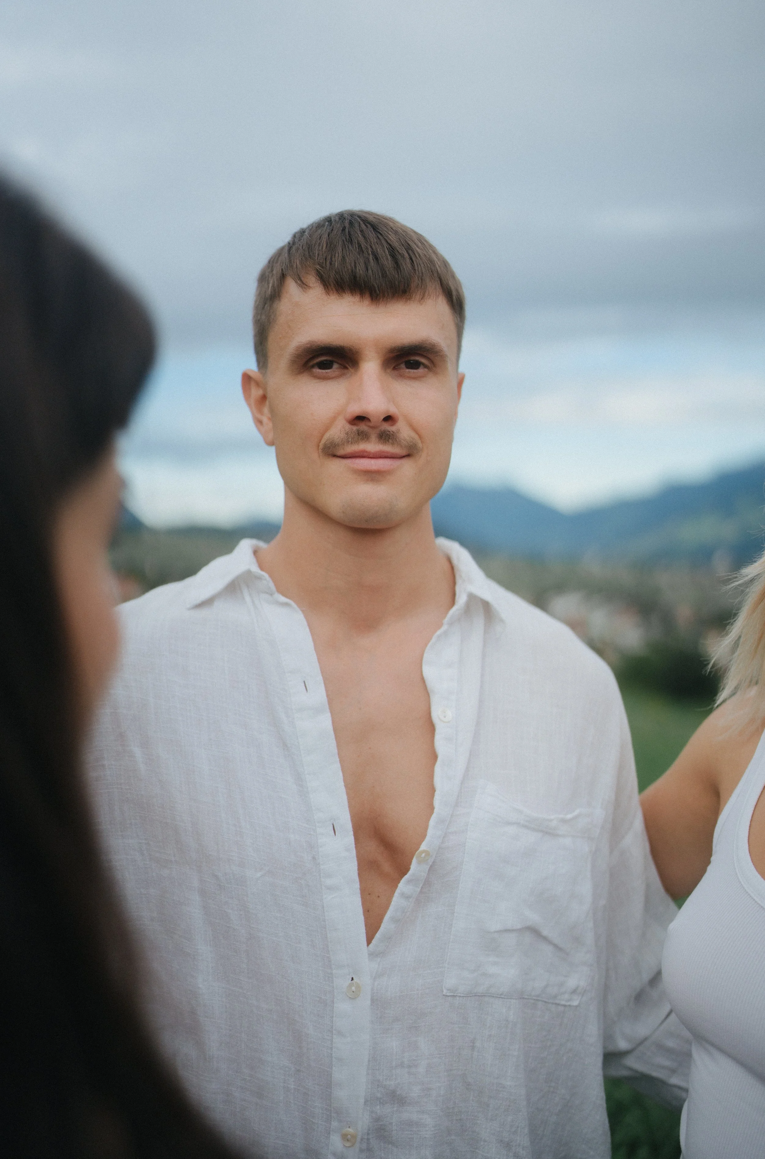 A young man with short brown hair, wearing a gray t-shirt and a blue denim jacket, smiling outdoors during daytime with blurred autumn trees in the background.