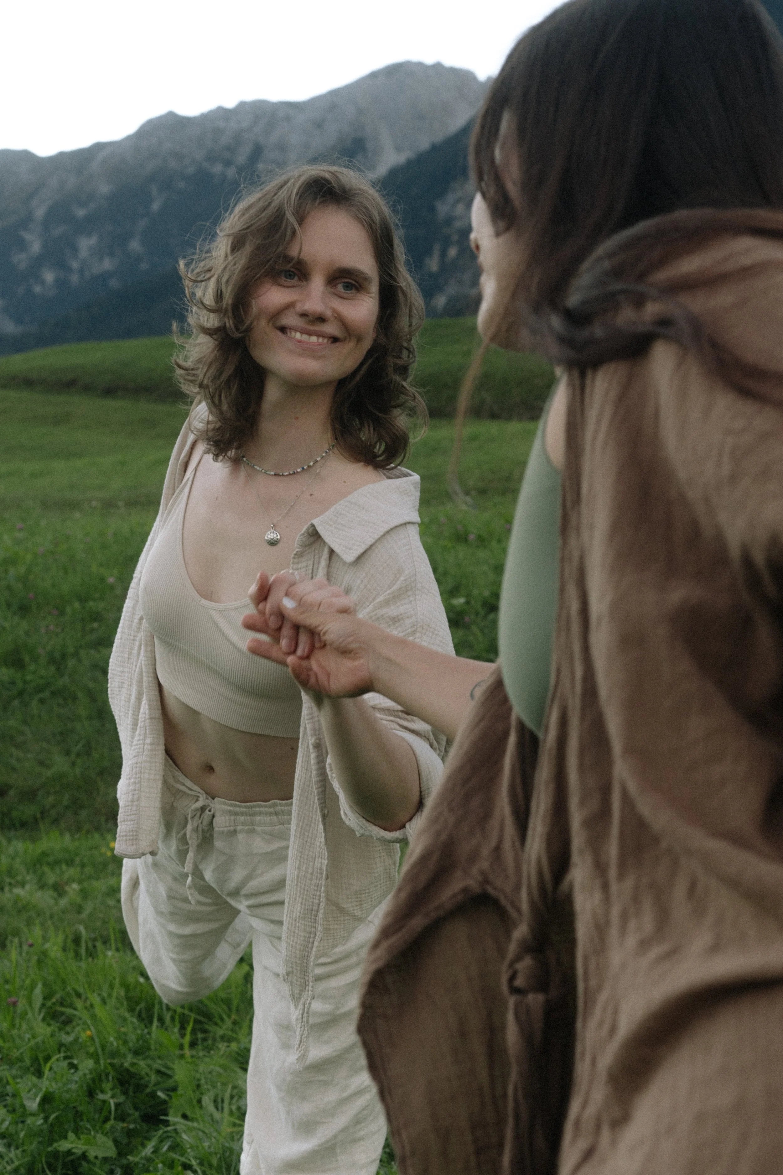 A woman with shoulder-length wavy brown hair, wearing a beige crocheted crop top, a gray cardigan, and beige pants, standing against a plain white wall, smiling at the camera.