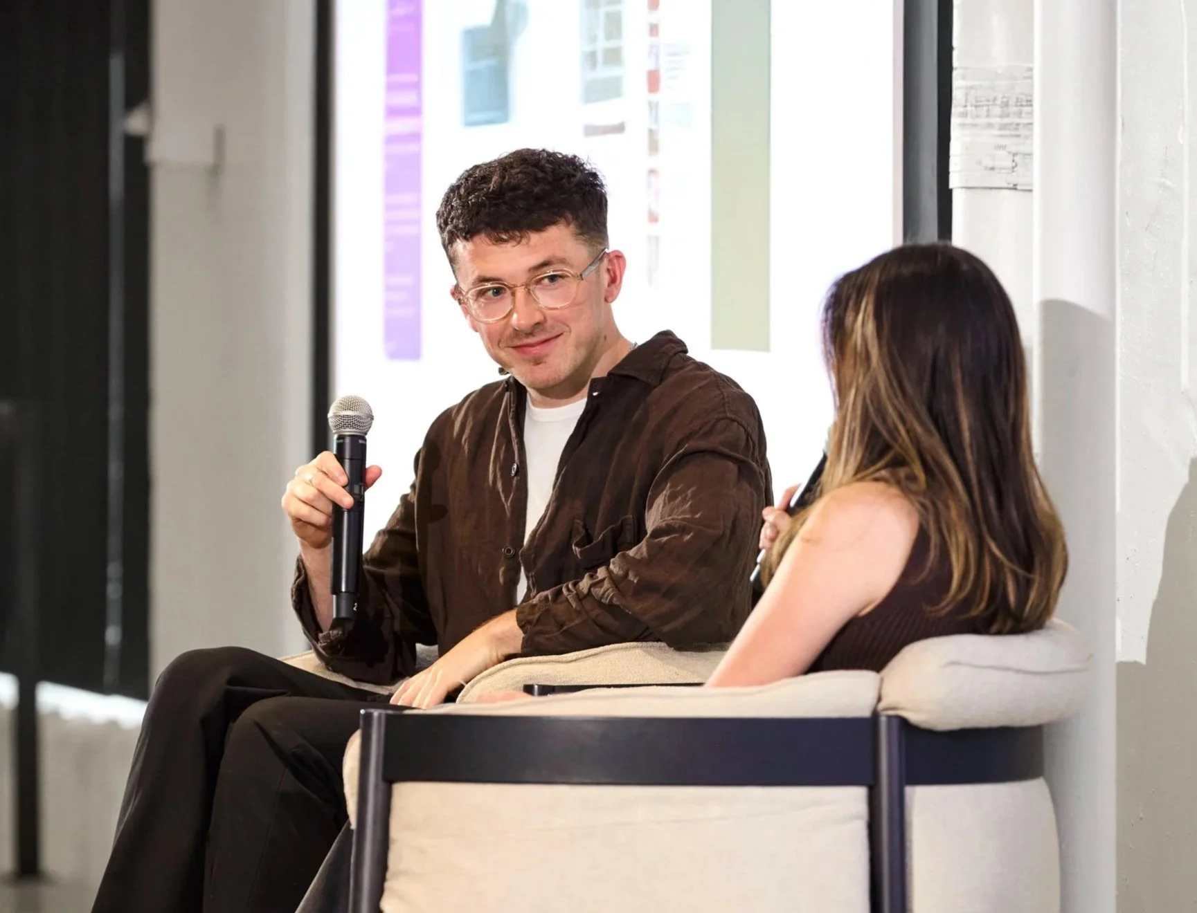 A man and a woman sitting on chairs and talking. The man is holding a microphone and looking at the woman, who is also holding a microphone. They are likely participating in a discussion or interview.