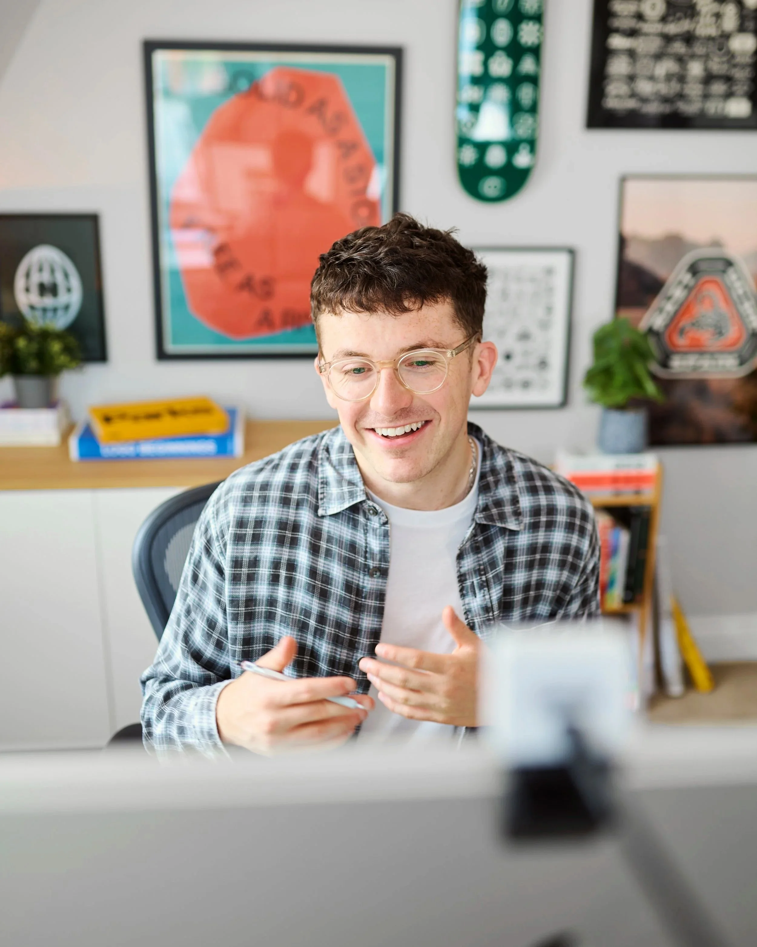 Young man with glasses smiling while on a video call or recording in a room decorated with framed posters and art.