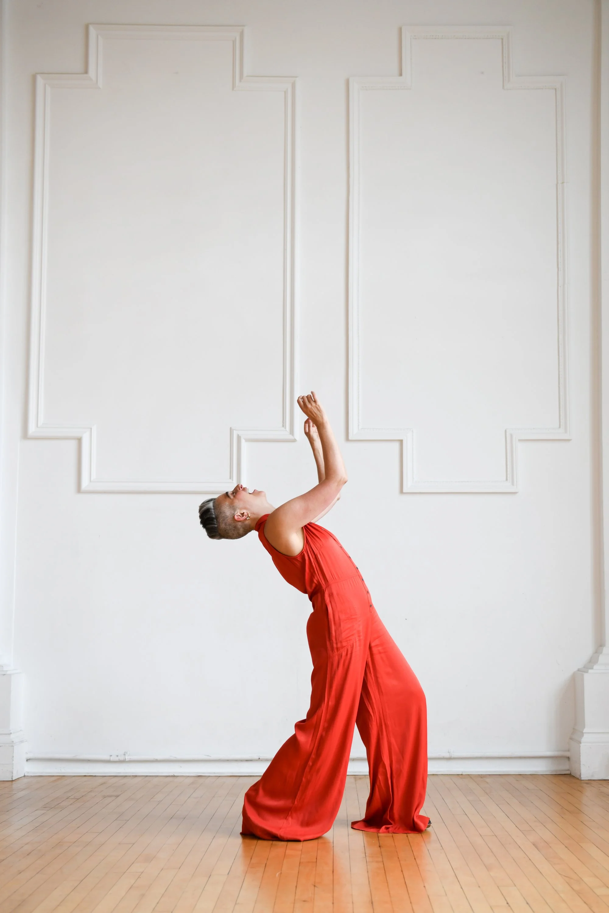 A person with short hair wearing a red sleeveless jumpsuit, standing in a dance pose in front of a white wall with decorative molding, on a wooden floor.