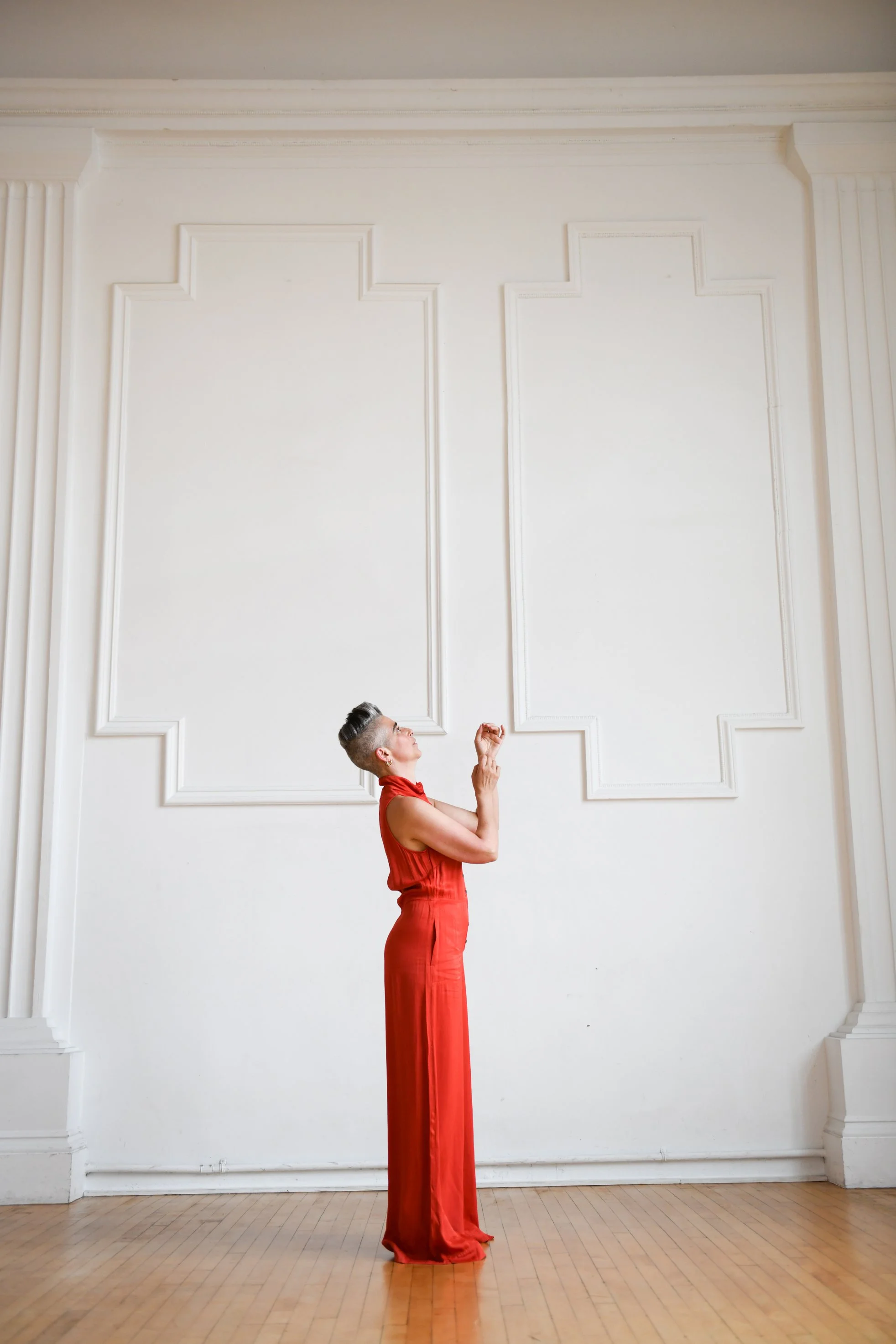 A woman in a long red dress stands on a wooden floor in front of a white wall with decorative molding, looking up with her hands raised.