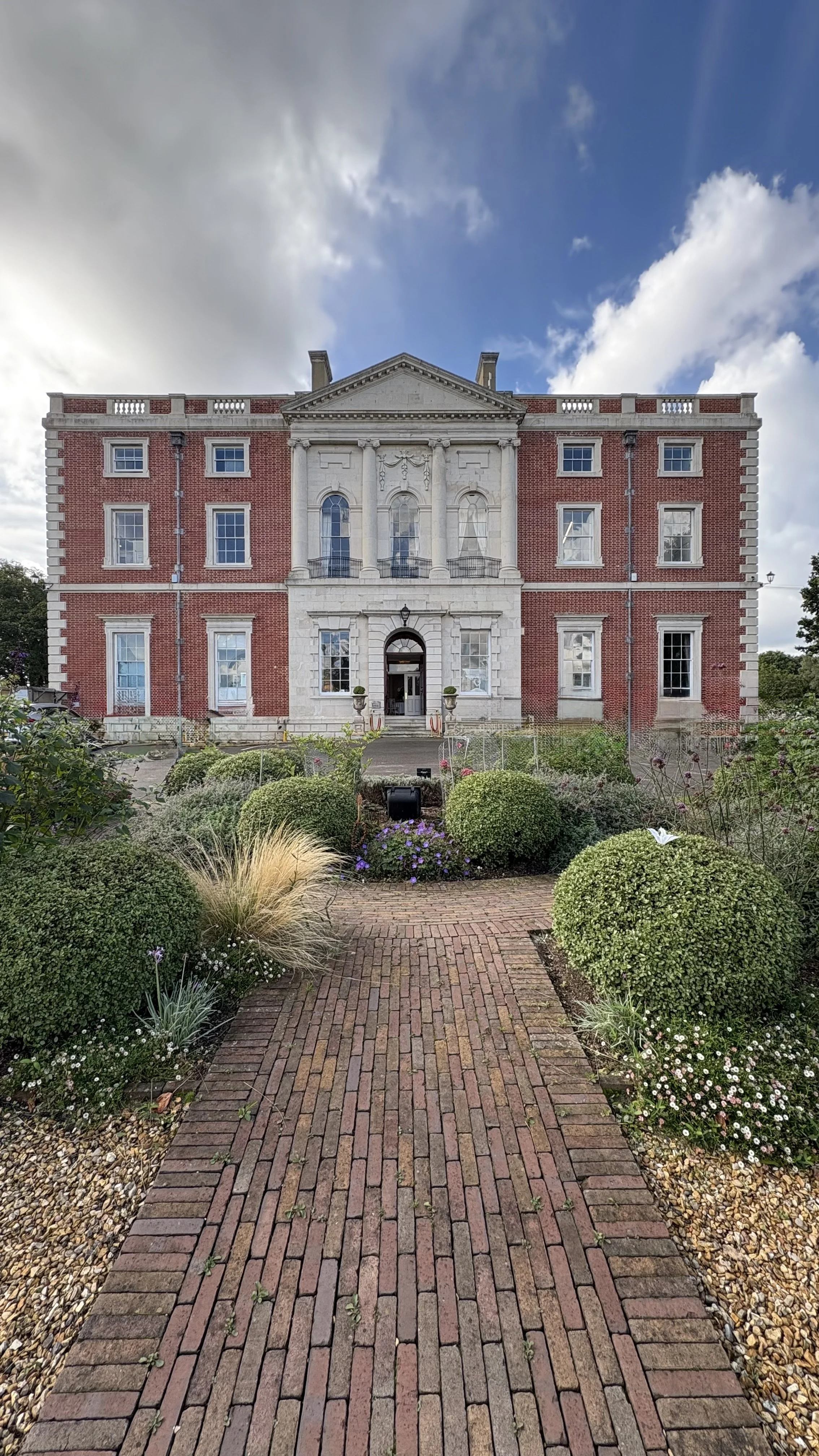 Large historic brick mansion with stone columns and a front garden with a brick path, bushes, and flowers.