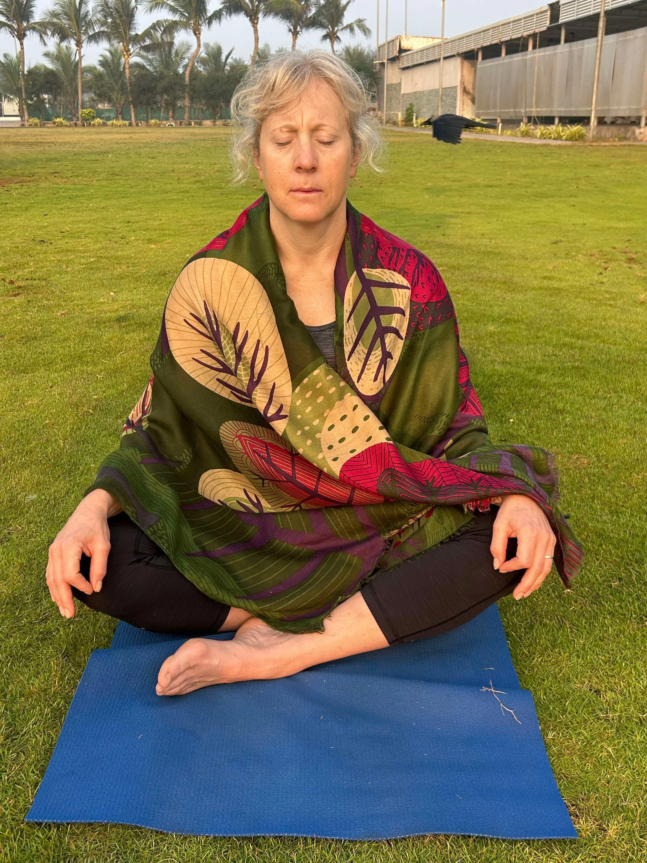 A woman practicing yoga outdoors on a green lawn, sitting cross-legged on a blue yoga mat with her eyes closed, wearing a colorful patterned shawl and black leggings, with palm trees and a building in the background.