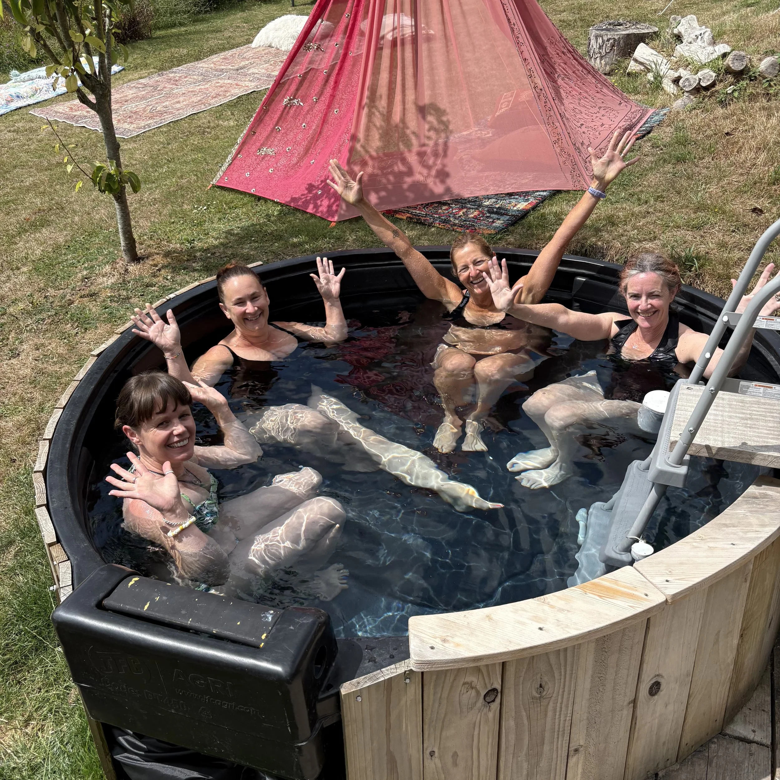 Four women sitting in a round hot tub outdoors, all smiling and waving. There is a pink canopy and blankets on the grass nearby.