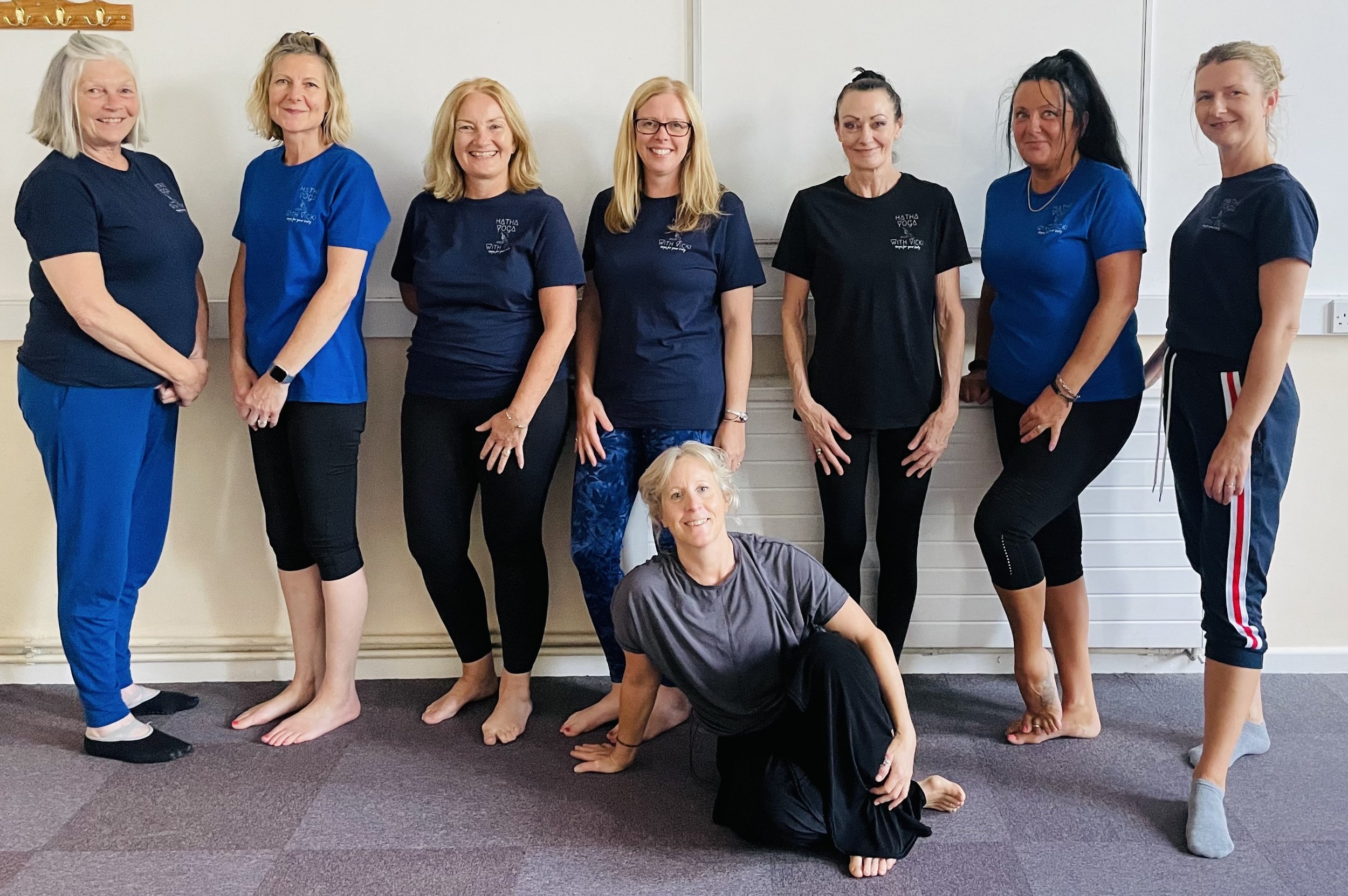 Group of eight women, seven standing and one sitting on the floor, in a classroom or studio with white walls, dressed in casual athletic clothing, some in matching blue shirts, smiling for the photo.