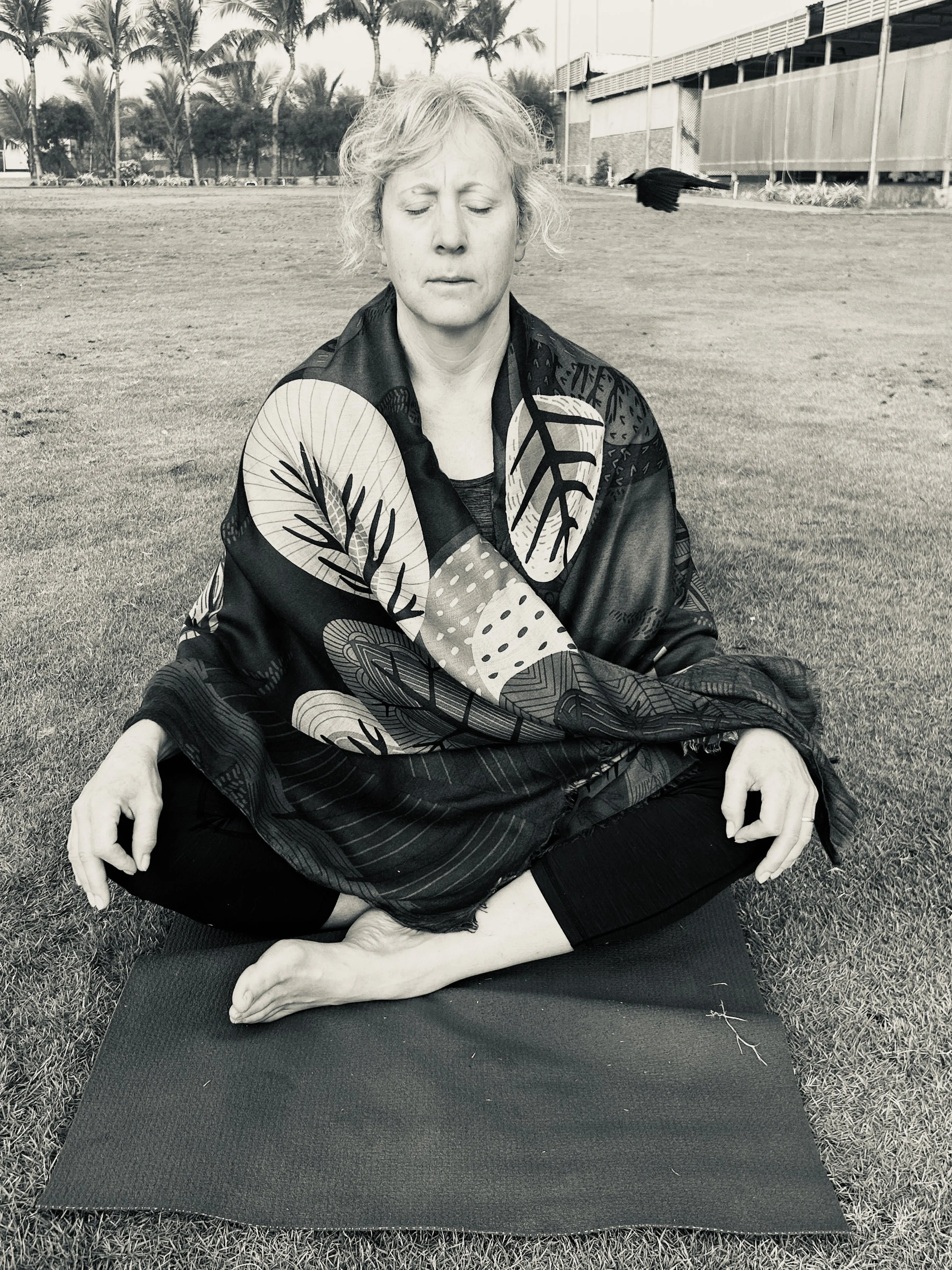 A woman practicing yoga outdoors on a mat, sitting cross-legged with her eyes closed and a serene expression, surrounded by a grassy field and palm trees.