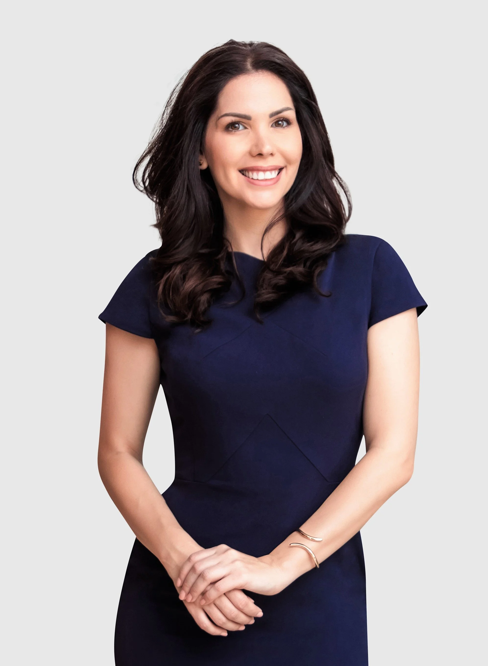 A woman with long dark hair, smiling, wearing a navy blue dress, standing with her hands clasped in front of her against a plain light gray background.