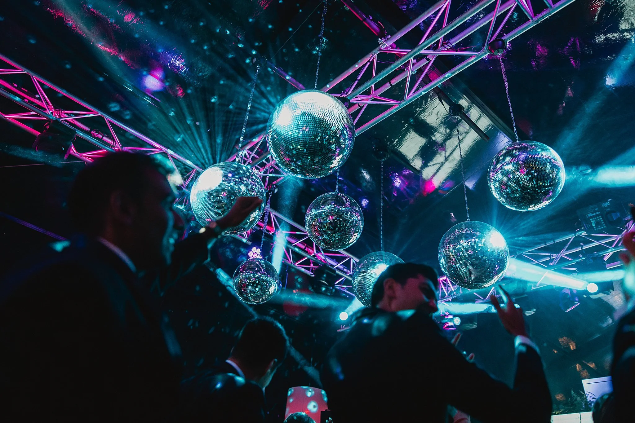 Multiple disco balls hanging from a ceiling with colorful purple, blue, and pink lights reflecting off them, creating a vibrant party atmosphere.