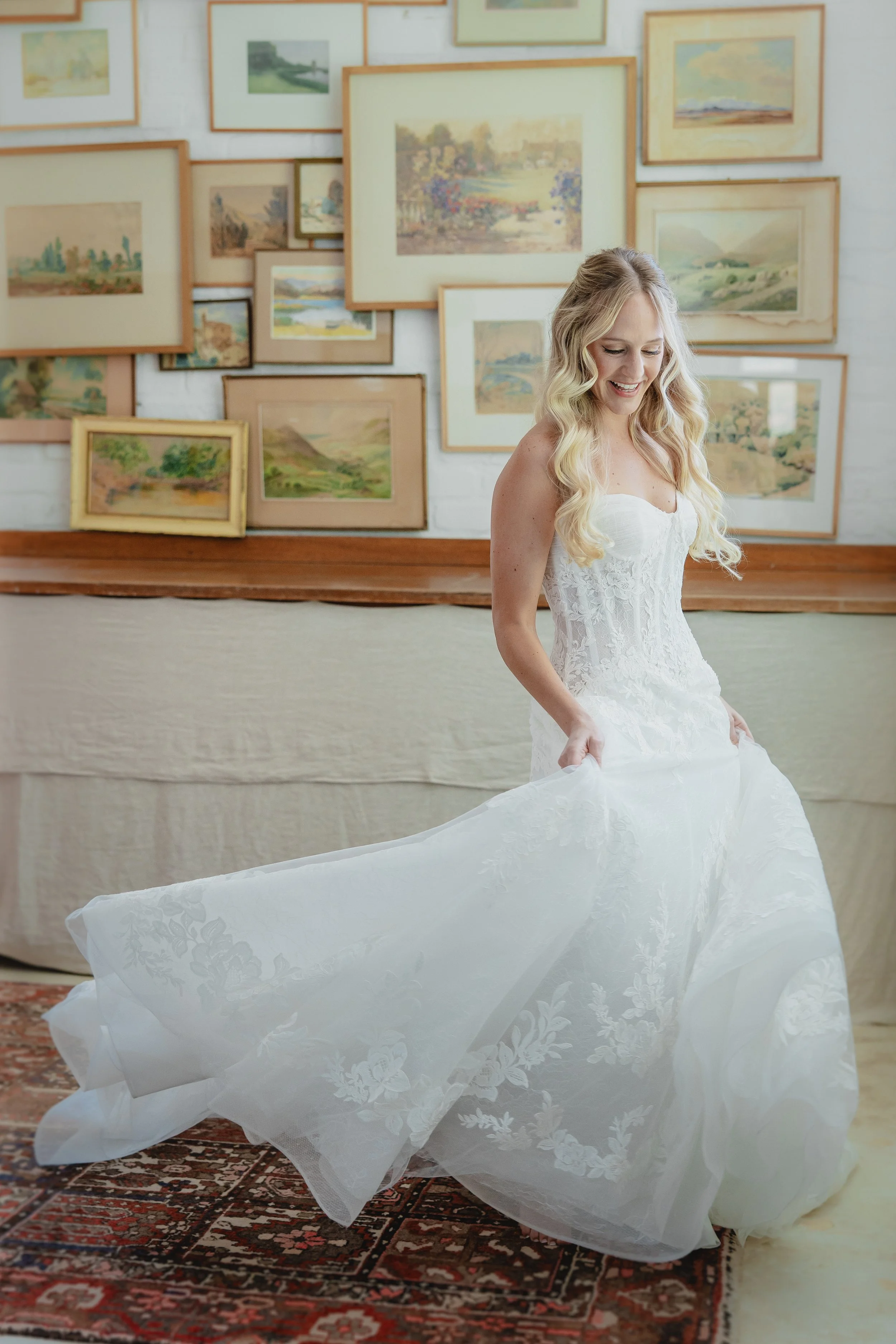 A smiling woman with long blonde hair in a white wedding dress, holding the full skirt of her dress, standing indoors in front of a wall decorated with multiple framed landscape paintings.