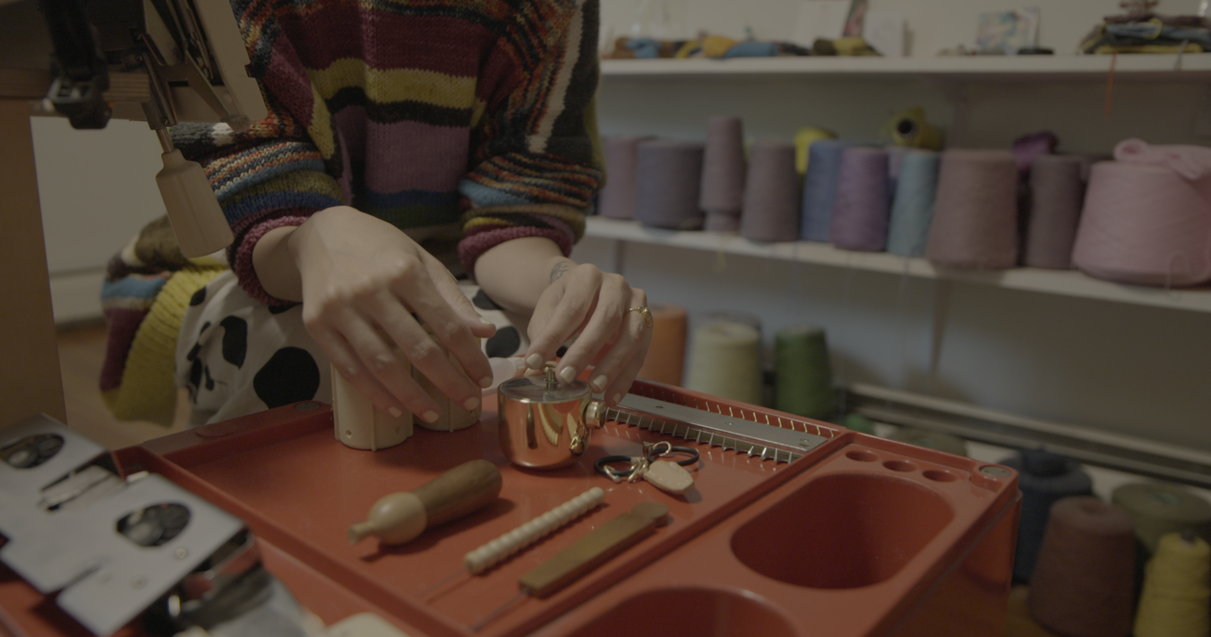 The Domestic Machine documentary still, close-up of hands adjusting a knitting machine's tension mechanism with yarn cones visible in background directed by Craftwork