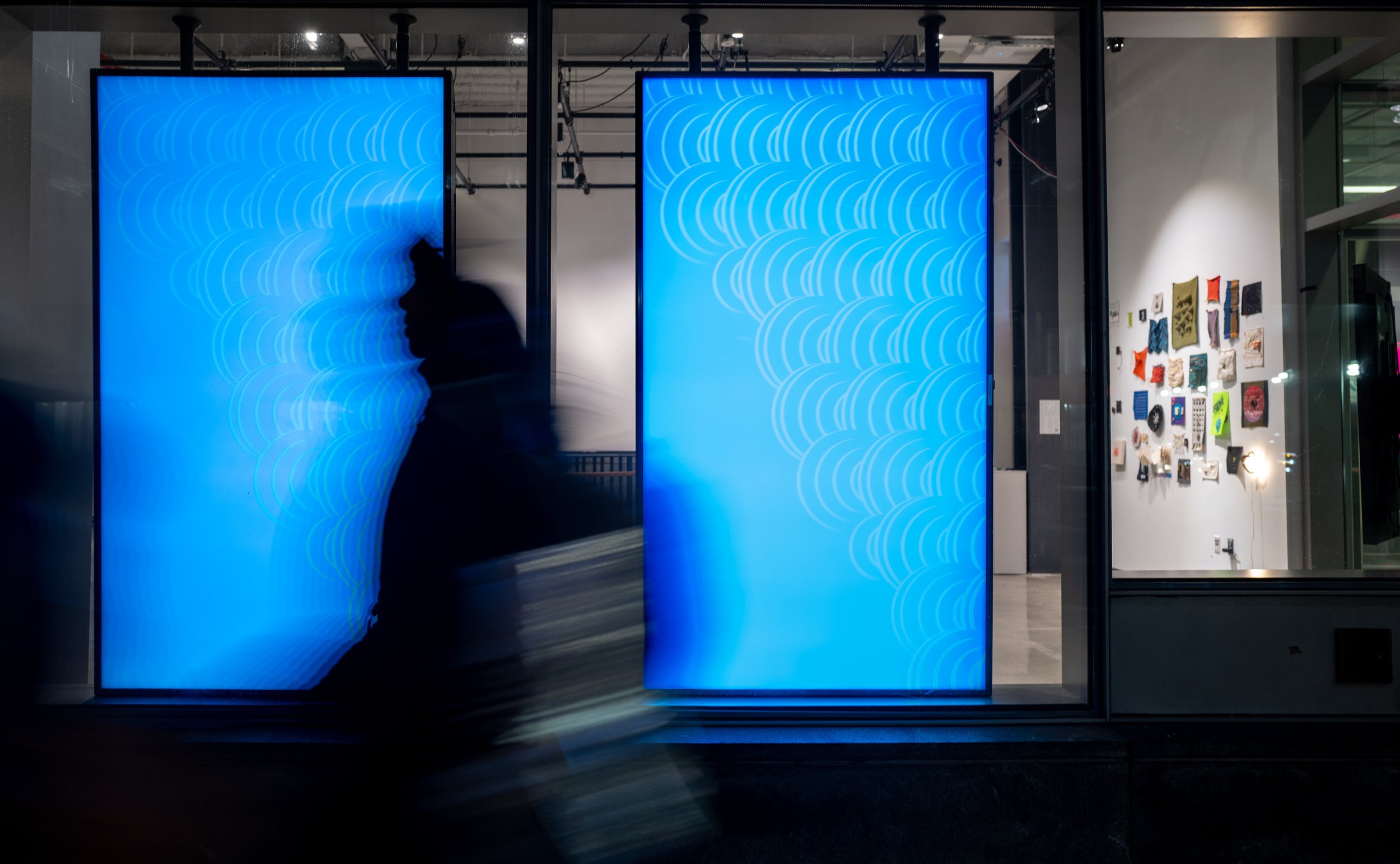 Two tall glowing blue screens in a storefront window at night show white arch patterns while a blurred figure walks past.