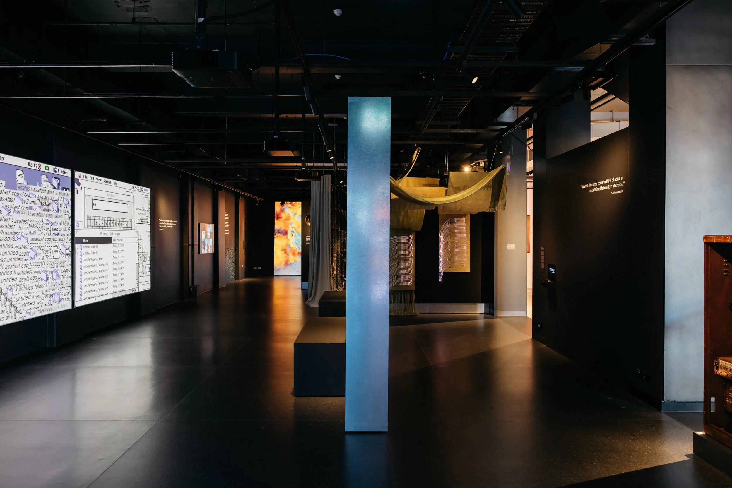 Craftwork Ancient Futures installation viewed from the Signal to Noise gallery corridor, illuminated woven panels visible beside vintage computer displays and quote wall