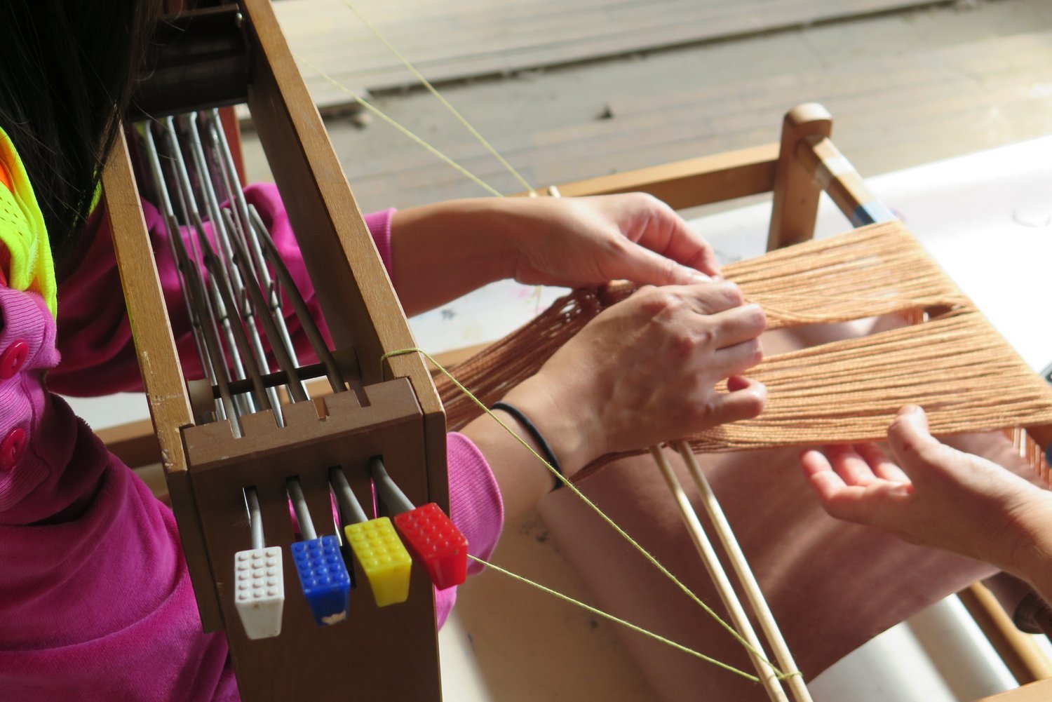 Hands manipulating colorful threads on a weaving loom with LEGO block color indicators, Electronic Textile Camp (ETC) co-organized by Craftwork