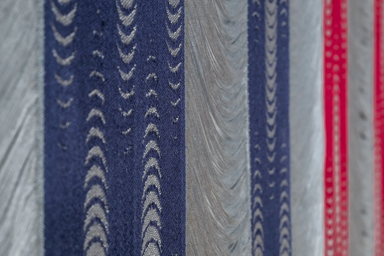 Close-up of navy woven bands with columns of pale gray crescents, beside draping gray threads and blurred crimson bands.