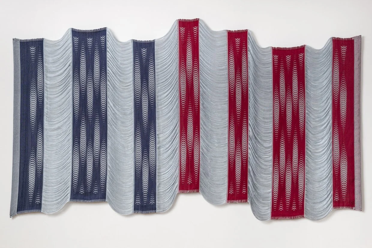 Wide wall textile of navy and crimson woven bands patterned with crescents, joined by draping pale gray threads.