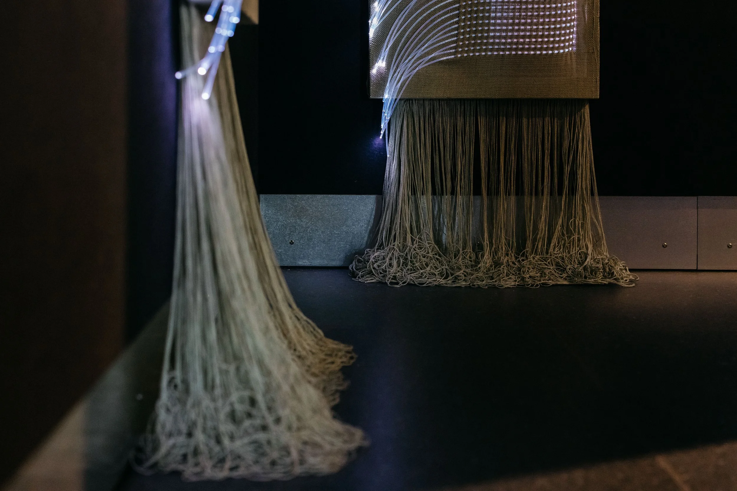 Craftwork Ancient Futures installation at NCM, fiber optic threads and unraveled fringe hanging from woven panels in a dark gallery
