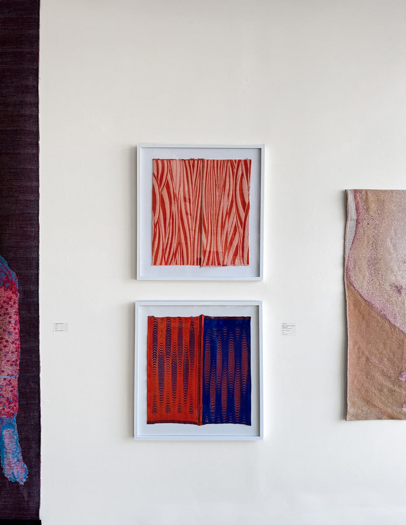 Gallery wall with two white-framed woven textiles stacked vertically: one orange and pale pink, one orange and cobalt blue.