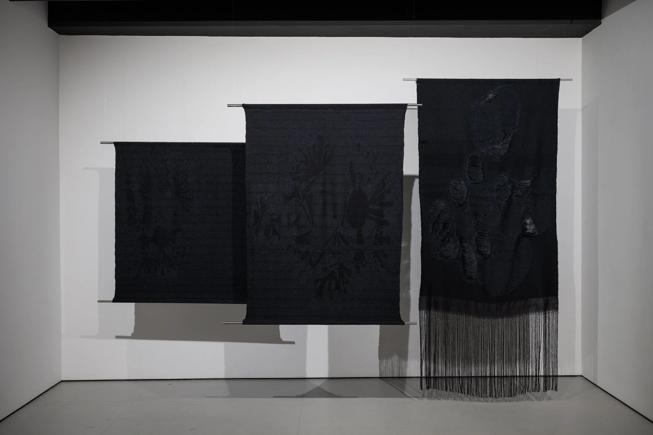 Slow Medium installation view, three dark woven textile panels of  varying heights with fringe detail, by Craftwork