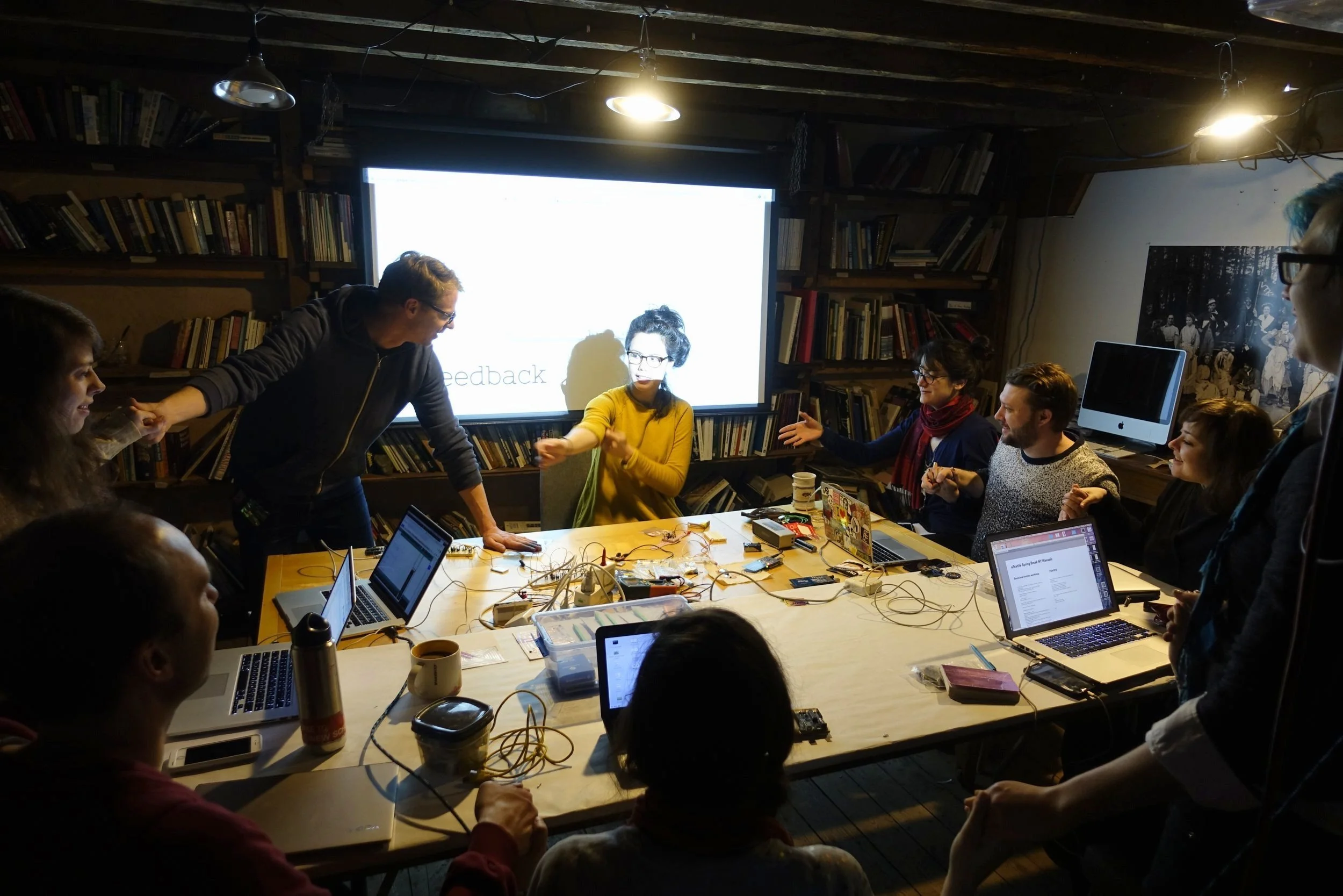 Group of participants working with laptops, electronics, and textiles around a workshop table with presentation screen, Electronic Textile Camp (ETC) co-organized by Craftwork
