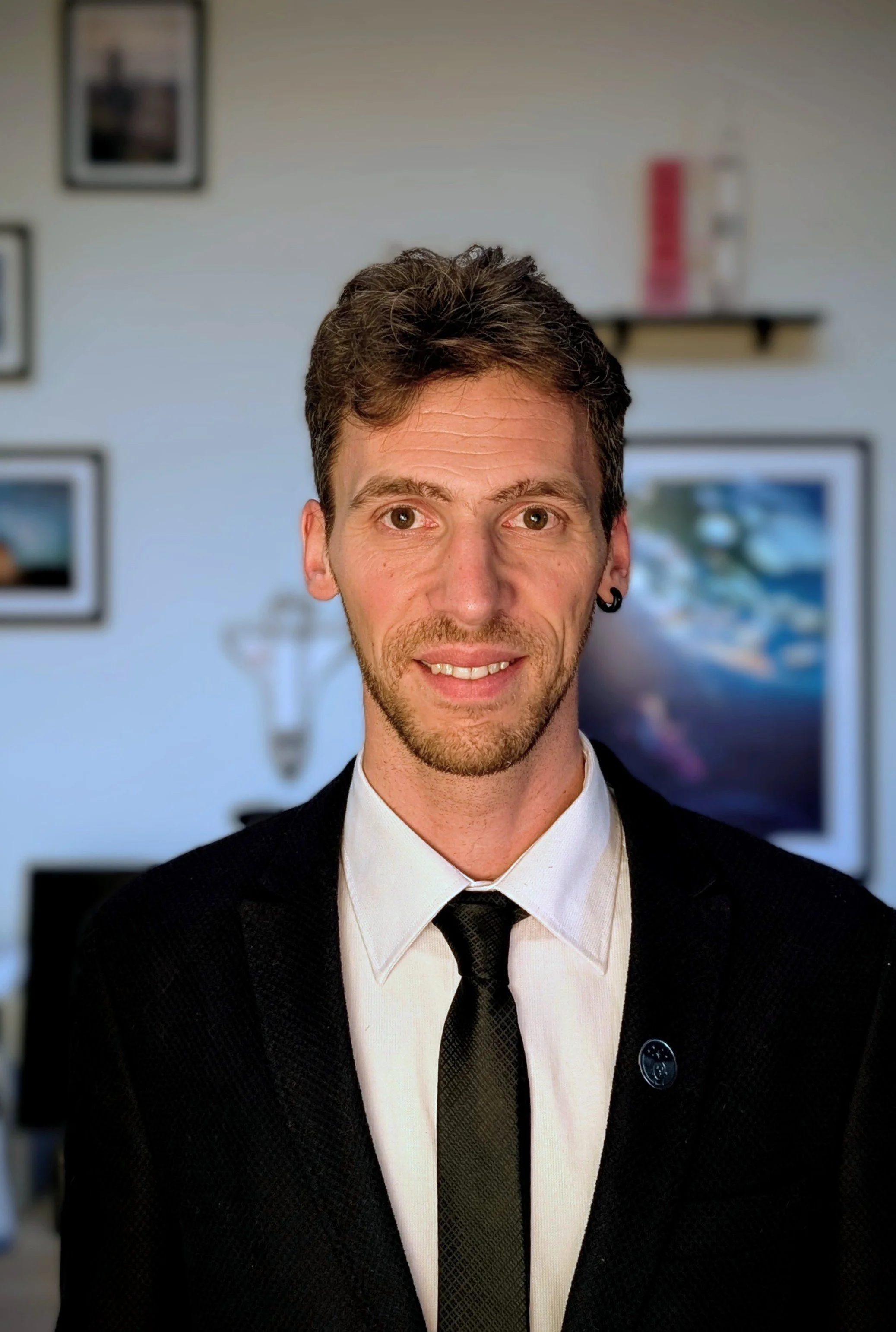 Retrato de un hombre con cabello castaño y barba, usando camisa blanca, corbata negra y traje negro, en un fondo de interior con fotos y decoración en la pared.