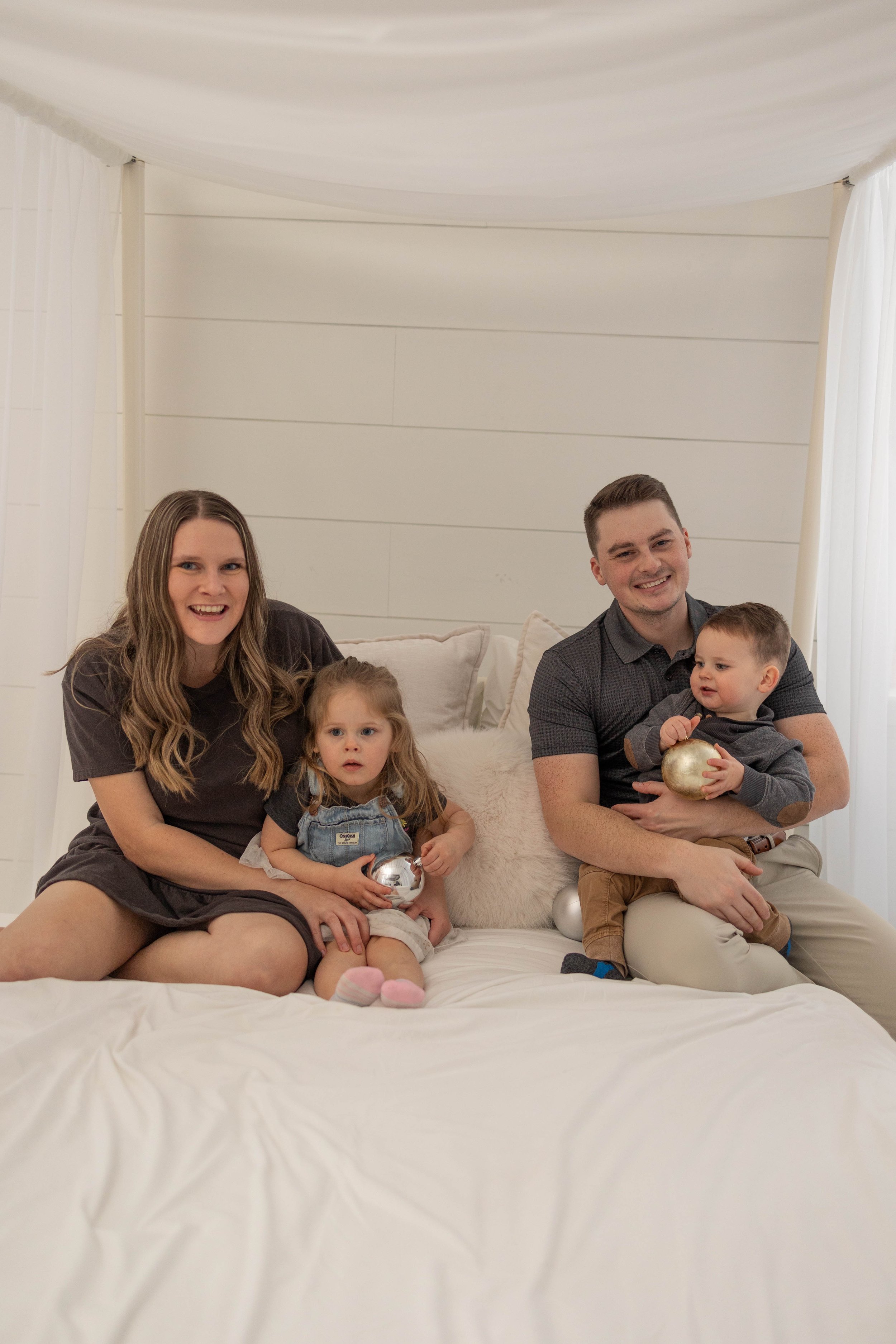 A family of four sitting on a bed in a cozy, well-lit room, smiling and holding Christmas ornaments.