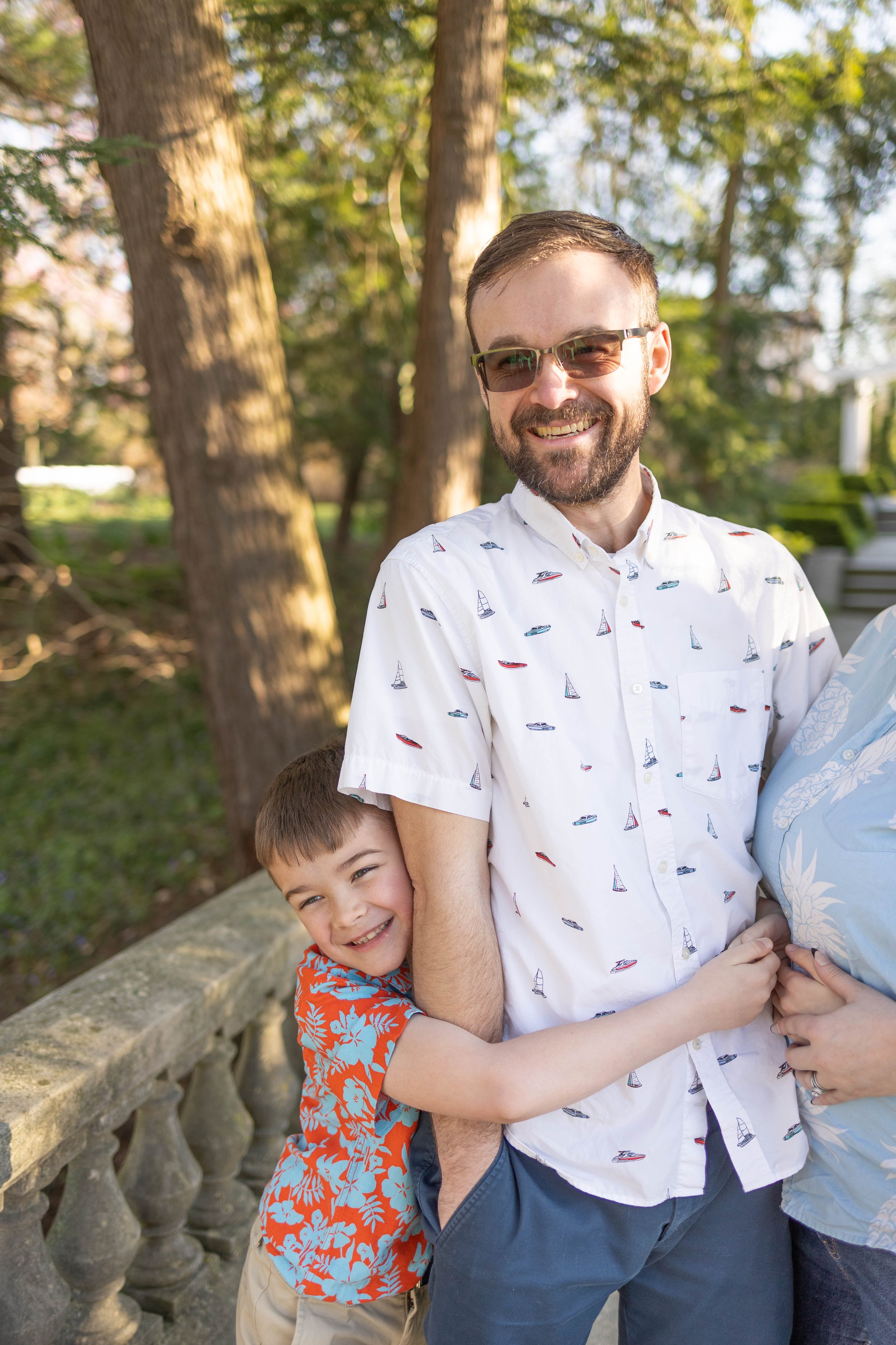 A man with glasses and a beard smiling, wearing a white shirt with sailboats, is hugging a young boy with brown hair, wearing a bright orange Hawaiian shirt, outside in a park with trees and sunlight.